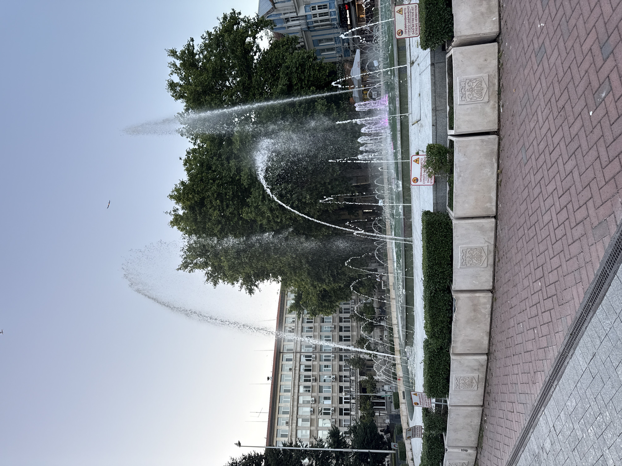 This is a medium shot, eye-level outdoor photograph of a large, multi-tiered fountain in a city square. The fountain features multiple streams of water shooting upwards, some with a wide spray and others as thin jets. In the background, there are tall, leafy green trees and several buildings. The building directly behind the fountain is a light-colored, multi-story structure with many windows. To the right, a building with blue and white striped facade is visible. The foreground consists of a red brick paved area, with a sidewalk of grey pavers to the left. Along the edge of the fountain are large, rectangular concrete planters containing green shrubs. Signs with warning symbols are posted on some of the planters. The sky is clear and light blue, with a few birds visible. The lighting suggests it is daytime, likely late afternoon or early morning due to the soft light. There are no people clearly visible in the immediate vicinity of the fountain, though figures can be seen in the distance on the sidewalk.