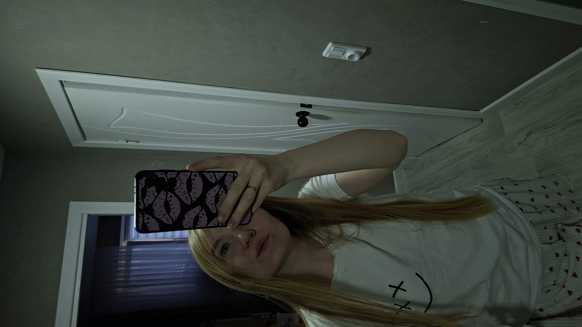 A young woman with long blonde hair is taking a mirror selfie in what appears to be a residential hallway. She is wearing a white t-shirt with a simple smiley face design and patterned shorts. Her right hand is holding a smartphone with a case featuring a pattern of purple lips. A white door with a black doorknob is visible behind her, to her right. The wall appears to be painted a neutral gray, and there is a light switch and outlet on the wall to the right of the door. The floor is covered in light-colored wood-look laminate. The lighting is dim, suggesting it may be evening or indoors with limited natural light. The overall impression is an informal, personal moment captured at home. There is no information in the image to suggest it is located in Chaikovsky, Russia.