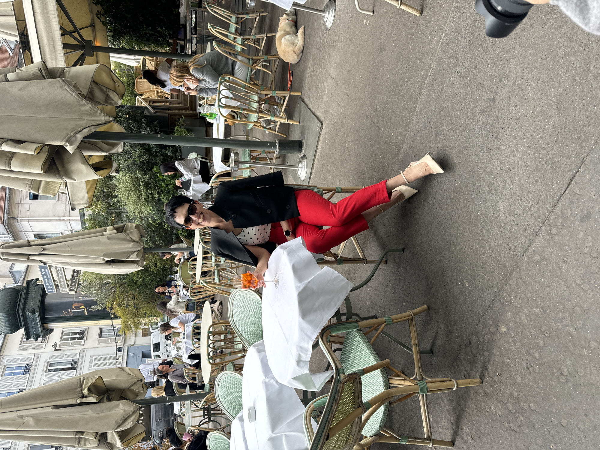This image captures a vibrant daytime scene at an outdoor cafe in Paris, France. The central subject is a stylish woman seated at a table, looking directly at the camera with a smile. She is wearing sunglasses, a black jacket over a white polka-dotted top, and bright red pants, with her legs extended. She holds a glass containing an orange drink. A hand and camera lens are visible in the upper right, indicating she is posing for a photograph.
The cafe area is furnished with numerous tables draped in white tablecloths and wicker chairs featuring light green accents, partially shaded by large, folded beige patio umbrellas. Other patrons are visible throughout the outdoor seating, some engaged in conversation or looking at their phones. A small, light-colored dog is seen curled up and sleeping under a table in the background.
The setting features typical Parisian architecture with multi-story, light-colored buildings and windows adorned with shutters. An ornate green lamppost or monument base stands on the left side of the frame. Crucially, a street sign in the background clearly reads "Bureau de Poste," "Jardin de Trocadéro," and "Palais de C," confirming the location in Paris, specifically near Trocadéro. The atmosphere appears relaxed and inviting, under natural daylight conditions.
