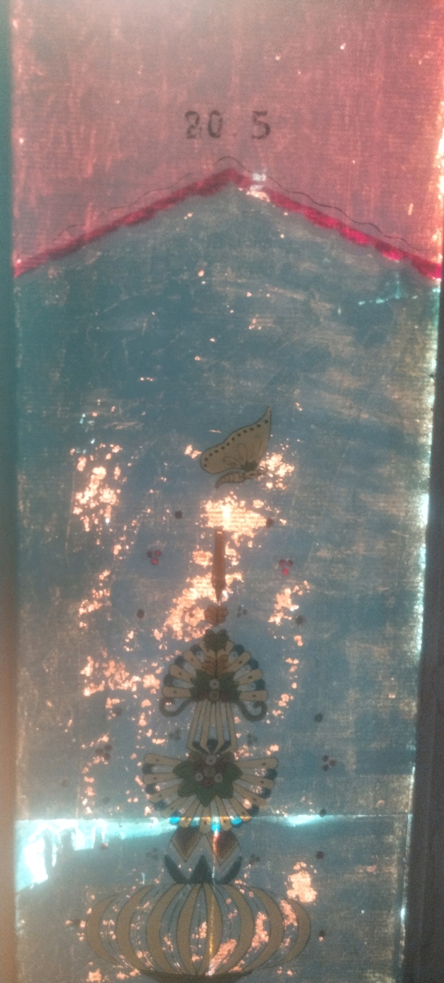 The image shows a close-up view of a stained glass window, possibly from a building in Daleman, Indonesia. The window features decorative elements, including what appears to be a butterfly motif and floral designs. The upper portion of the window has the numbers "20.5" printed or etched onto it. The glass is somewhat aged and shows signs of wear, with some discoloration and scattered light-colored flecks. The background is a mottled blue, with a reddish-pink border at the very top.