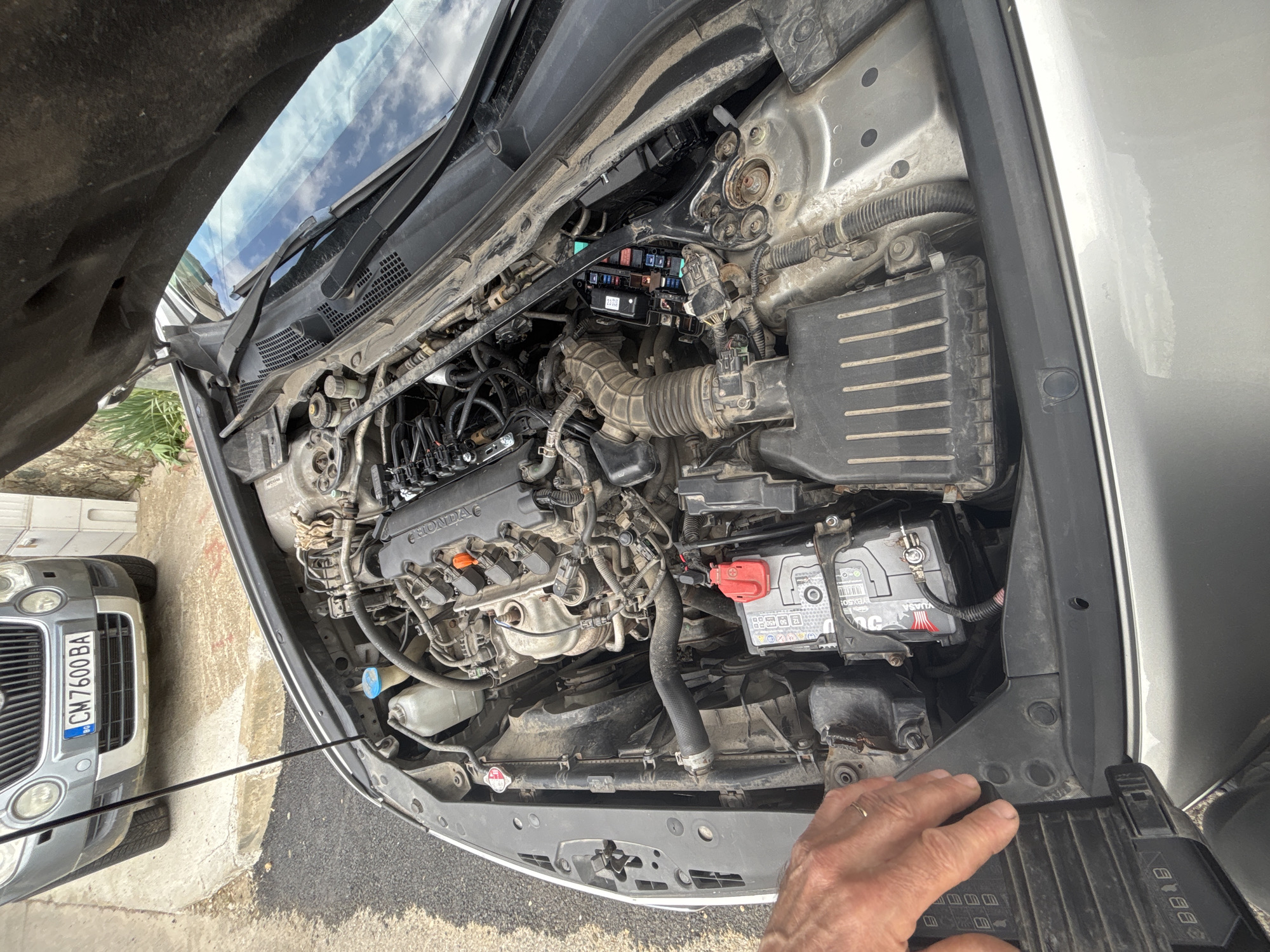 The image shows the open hood of a silver Honda car. The engine is visible with various components such as the engine block, air intake, battery, and fuse box. A hand is visible near the front of the car, holding a black part. Another silver car is partially visible in the bottom left corner of the image, with a license plate that reads "CM 7600 BA". The location is likely outdoors on a paved surface. Based on the license plate, the location may be in Bulgaria.