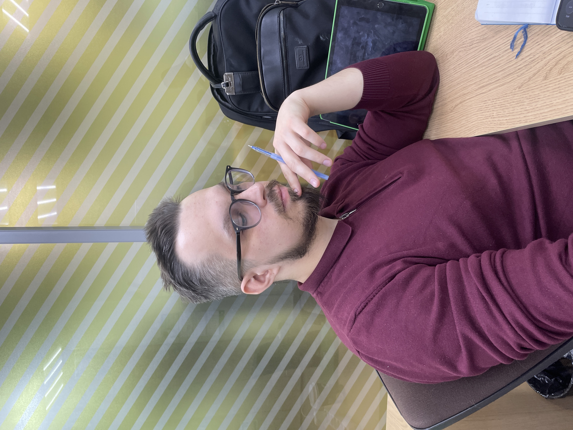 An overhead, slightly angled shot depicts a man seated at a wooden table in an indoor setting. The man, with a neatly trimmed beard and mustache, wears black-rimmed glasses and a maroon-colored sweater. He appears pensive, holding a blue pen to his chin with his right hand, and gazing upwards and to his left.
On the wooden table, to the man's right, rests a black backpack with silver hardware, including a small rectangular metallic badge that displays what looks like "CD" or "CP" intertwined, with additional faint text beneath it, possibly "Collection". Adjacent to the backpack is a tablet in a vibrant green case, its screen dark. Further right, a light-colored notebook with a blue bookmark is open on the table, showing some illegible writing.
The background features a wall adorned with a distinctive pattern of alternating light green and white diagonal stripes. The overall impression is one of quiet contemplation or study in what appears to be an office or educational environment. No specific time of day or weather can be discerned.