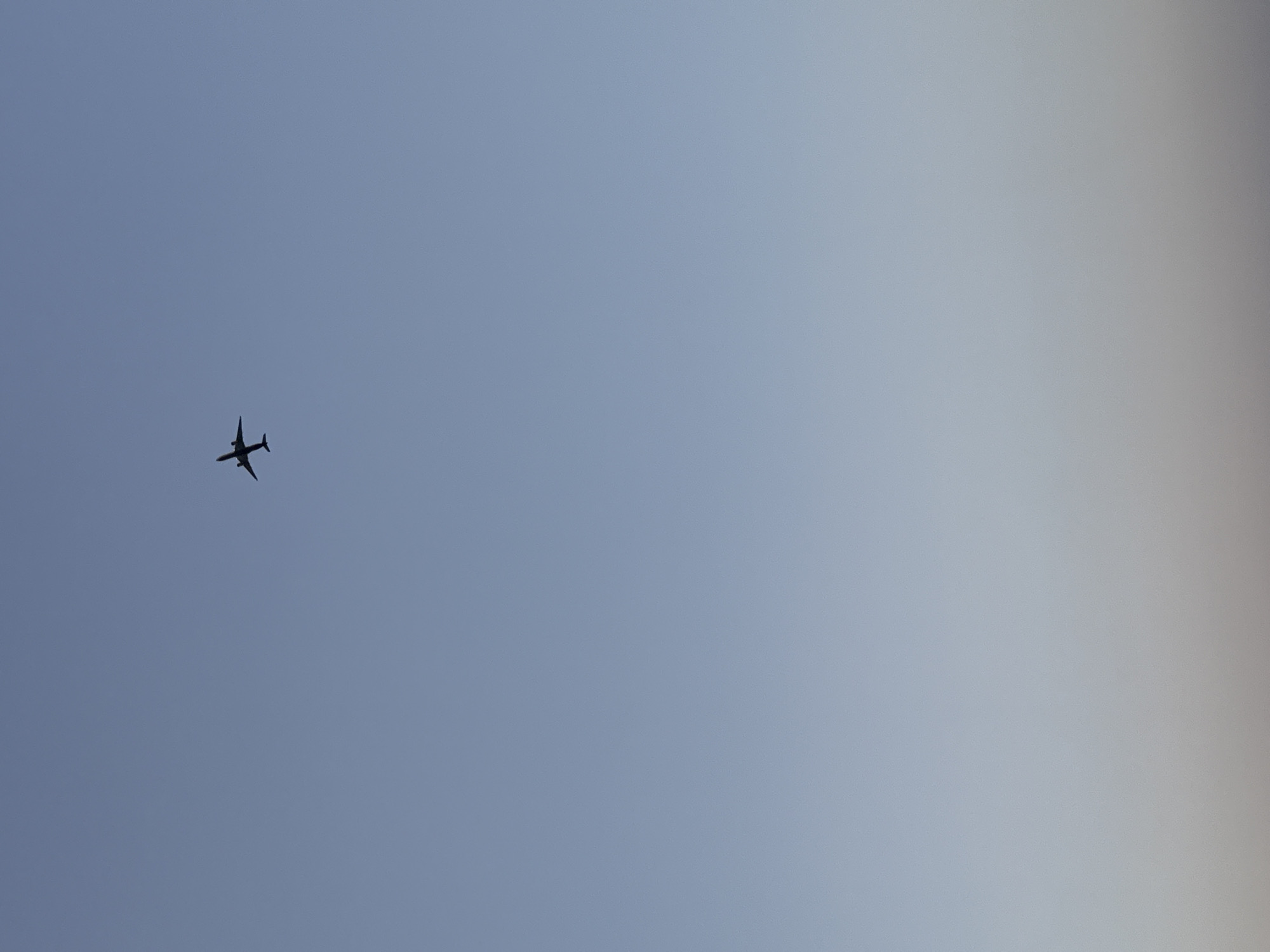 A silhouetted airplane is captured in flight against a gradient of soft blue sky, transitioning to a lighter hue near the horizon. The aircraft is positioned in the upper portion of the frame, slightly to the left of center. The image appears to be taken from the ground looking upwards. The location is the city of Castelldefels, Spain.