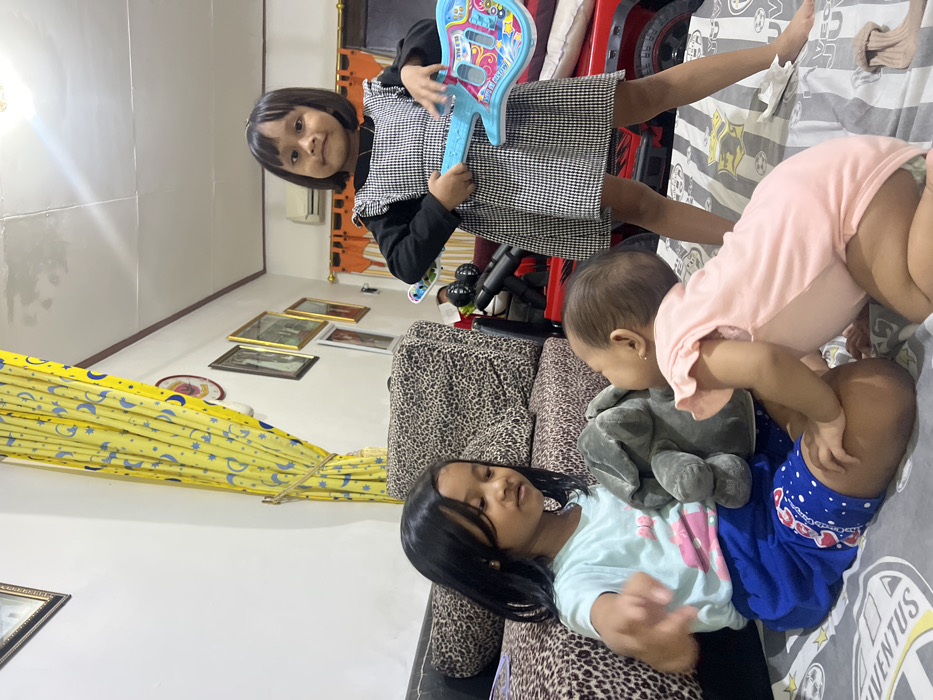 A domestic indoor scene in Telukan, Indonesia, depicts three children. A girl with dark, shoulder-length hair is seated on a gray and white patterned blanket, wearing a light blue short-sleeved top and dark blue shorts. She holds a gray plush elephant toy in front of her. Facing her, an infant wearing a light pink short-sleeved onesie is on all fours, partially obscuring the seated girl's lower body. To the right, another girl with short, dark bobbed hair stands barefoot on the patterned blanket. She wears a black long-sleeved top under a black and white gingham patterned sleeveless dress and holds a light blue toy guitar adorned with colorful decals. The background features a leopard-print upholstered sofa on the left, a white wall with multiple framed pictures, yellow curtains with blue moon and star patterns, and a partial view of a red toy car and orange curtains on the right. Overhead, a ceiling light fixture and an air conditioning unit are visible.