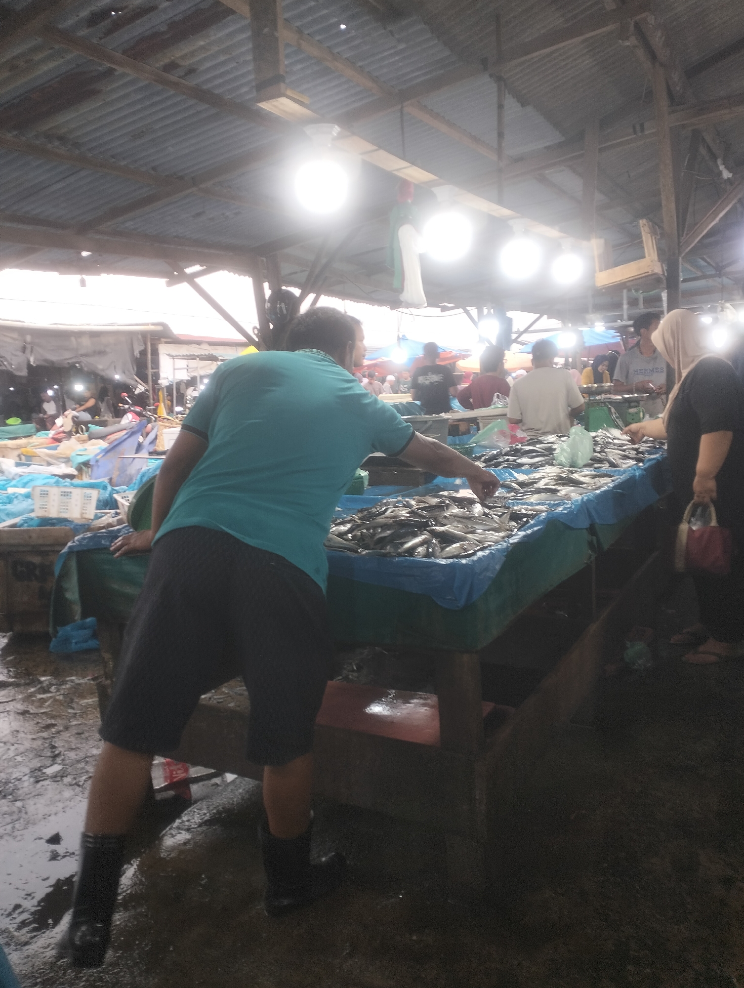 In Dumai City, Indonesia, a busy fish market is depicted under a corrugated metal roof supported by wooden beams. The market floor is wet and appears to be a mix of concrete and packed dirt. In the foreground, a person wearing a teal t-shirt and black shorts with black rubber boots is leaning over a table laden with fresh fish, their hand reaching out to select a fish. The table is covered with a blue plastic sheet and a green cloth. Behind this individual, several other people are visible, some tending to other stalls. Some stalls are covered with colorful tarps. The lighting is bright, coming from overhead bulbs, creating some glare and washing out details in the background. The overall scene suggests a bustling, active marketplace where fresh seafood is being sold.