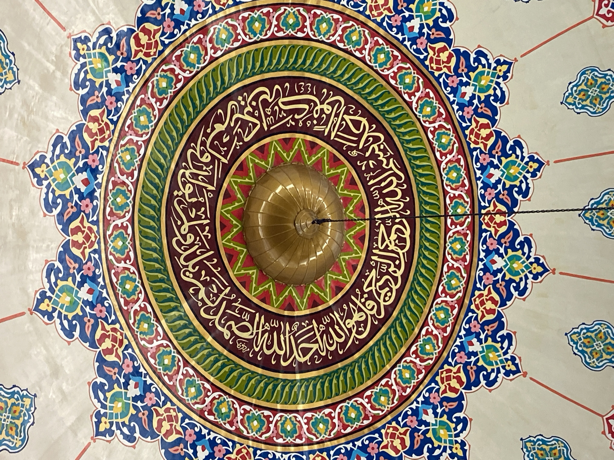 The image shows a highly ornate ceiling fresco in a building that appears to be a mosque. The fresco is circular, with intricate patterns in vibrant colors including blue, red, green, yellow, and gold. Calligraphy in Arabic script is prominently featured in gold against a dark red background in the central part of the circle. The center of the fresco has a golden dome, possibly a decorative element of the ceiling itself. The surrounding ceiling is a light, neutral color and decorated with smaller, repeating floral or geometric motifs and radiating red lines. The image is taken from a low angle, looking directly up at the ceiling.
The specific location of this ceiling fresco in Lhokseumawe, Indonesia, is not identifiable from the image alone. However, it is consistent with the decorative styles found in some Islamic religious buildings in Indonesia.
