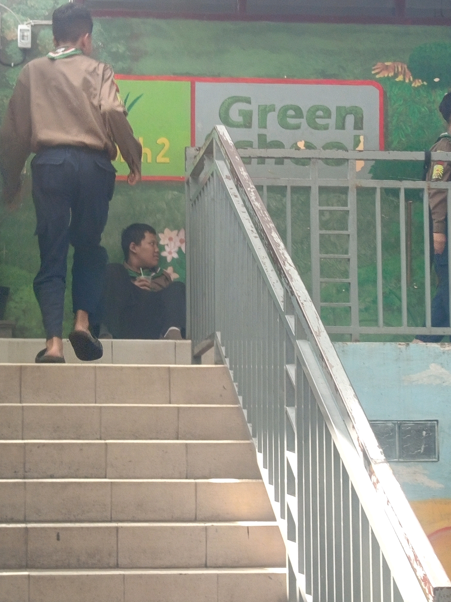 In the city of Malang, Indonesia, two individuals are present on an outdoor staircase. The person in the foreground, identified as a young male wearing a brown scout uniform and dark pants, is walking up the stairs. To the left and slightly behind him, another male is seated on the stairs, dressed in a black outfit and holding a cup with a straw. The background features a green mural depicting foliage, with a sign that reads "Green School 2." A metal railing runs along the side of the stairs, separating them from the mural. The lighting suggests it is daytime, with shadows indicating the sun's position.