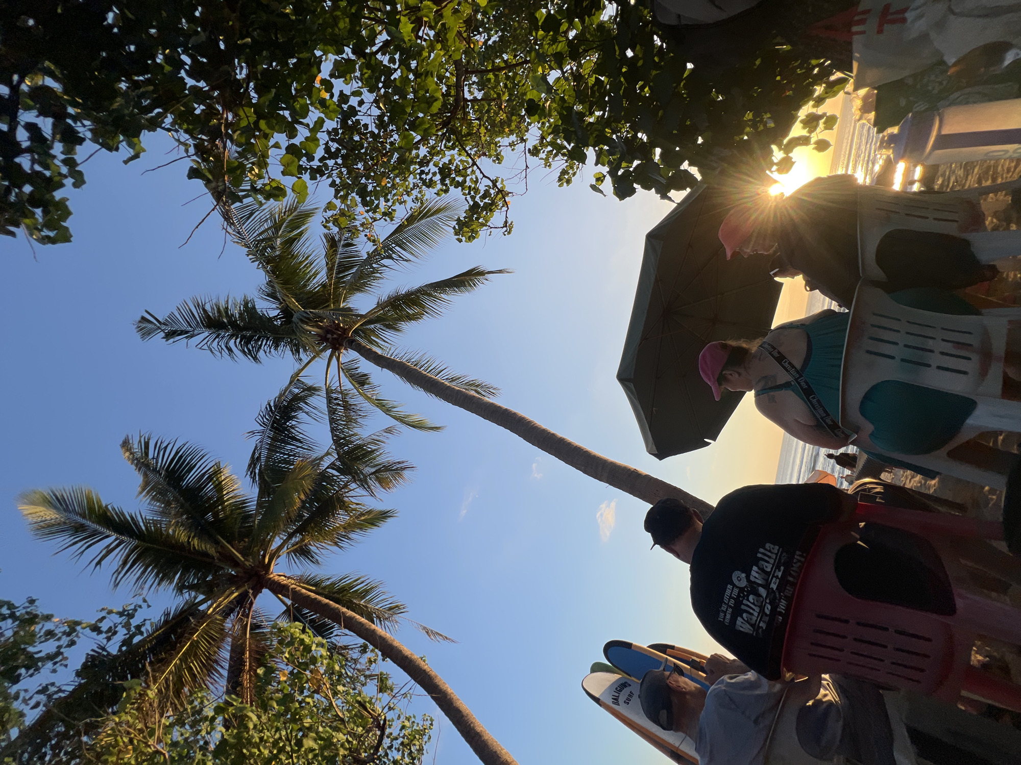 At sunset on Kuta Beach, Indonesia, three individuals are seated on chairs facing the ocean. Two of them are obscured by an umbrella, but the third, wearing a pink hat and a turquoise dress, is visible. To their left, two surfboards stand upright. The sky is clear and blue, with the sun setting at a low angle, casting a golden light and creating lens flares. Palm trees and foliage frame the scene, adding to the tropical ambiance.