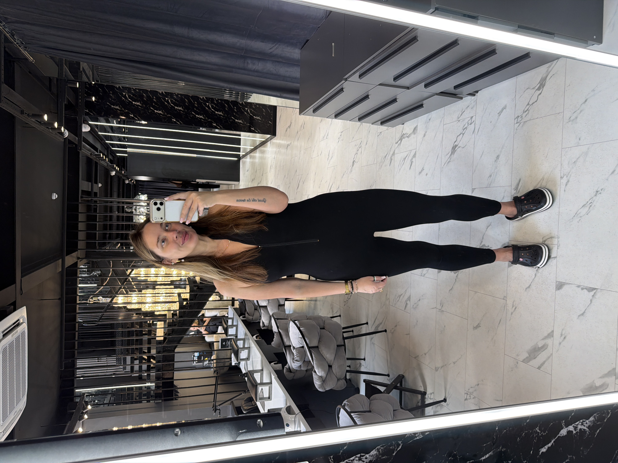 A woman with long brown hair stands in a salon or spa, taking a mirror selfie. She is wearing a black, sleeveless, zippered jumpsuit and black Crocs. The background shows a modern interior with marble flooring, black curtains, and a contemporary staircase with ornate lighting. To her right, there is a gray dresser with multiple drawers. In the reflection of the mirror, other salon chairs and workstations are visible. The woman is holding a white smartphone in her right hand. The location is identified as Укромновское сельское поселение, Ukraine.