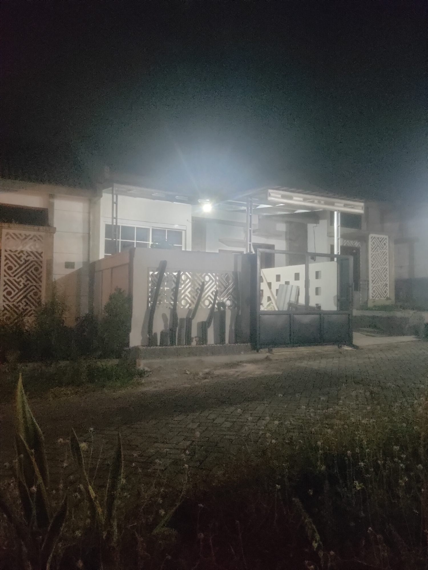 The image shows a building at night, with lights illuminating the facade. The building appears to be a house or a commercial property, with a modern design. There is a paved driveway in front of the building, and some greenery on the sides. The sky is dark, indicating that it is nighttime. The overall impression is one of a well-maintained property in a quiet neighborhood. The image is taken from a slightly elevated angle, looking towards the building. There are no people visible in the image.