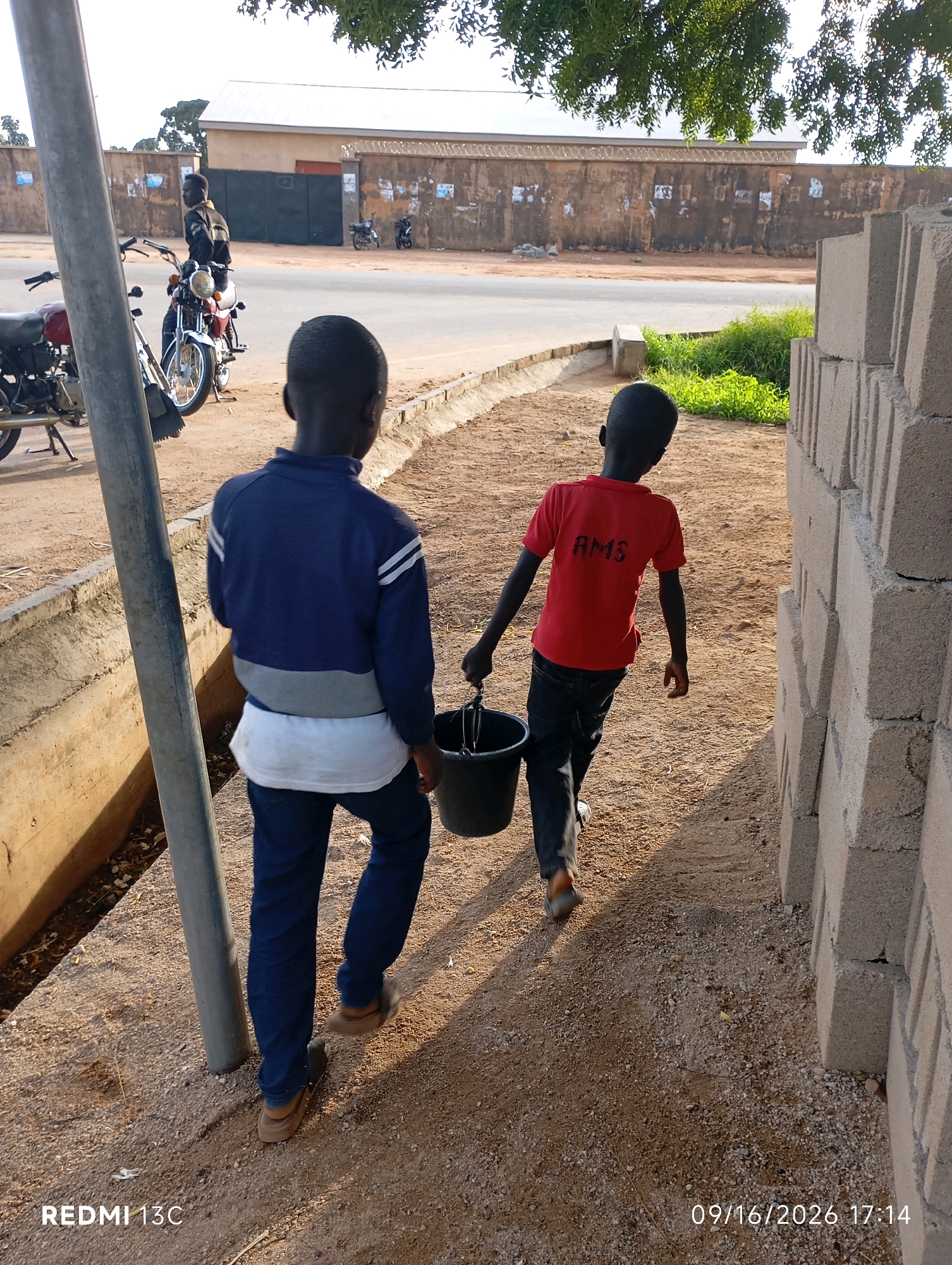 Two young boys are walking away from the camera on a dirt path. The boy on the left is wearing a blue jacket and blue pants. The boy on the right is wearing a red t-shirt with "AMS" printed on the back and black pants. He is carrying a black bucket in his right hand. To the right of the boys, a wall made of concrete blocks is under construction. In the background, a street is visible, with a man sitting on a motorcycle to the left. A building with a white roof and a beige wall stands behind the street. The environment appears to be an outdoor urban setting, possibly in Shendam, Nigeria, given the timestamps and context. The lighting suggests it is late afternoon.