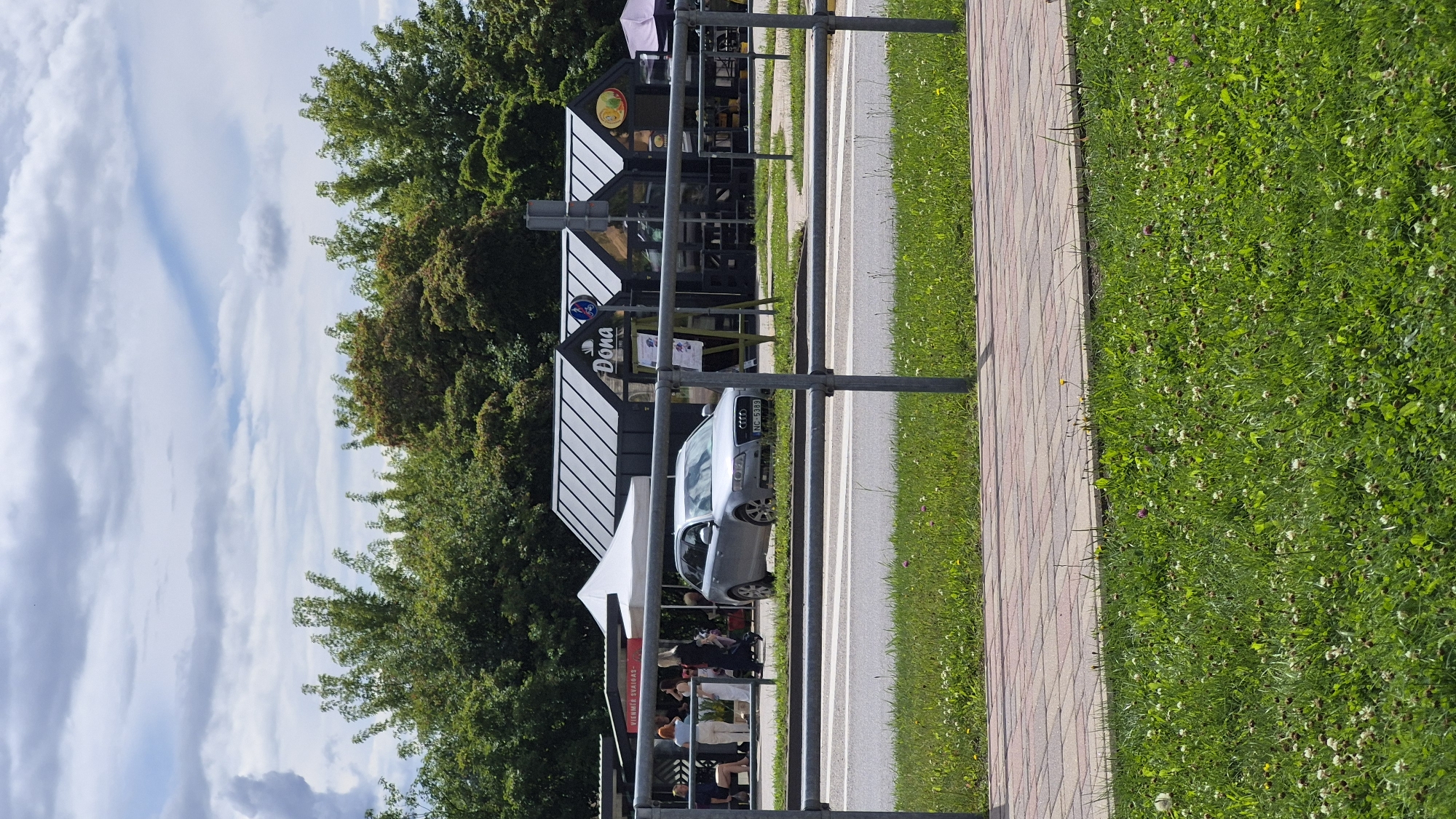 A silver Audi A4 sedan, license plate NC-5389, is parked in front of a modern-style, dark-colored building with a sloped roof. The building has the name "Dona" displayed prominently. To the left of the building, there is a white tent setup, and several people are standing near it. One person is seen pushing a stroller. In the background, lush green trees form a dense canopy. The scene appears to be outdoors, possibly a commercial area or a street in Jēkabpils, Latvia, given the presence of the distinctive building and the overall environment. The foreground shows a grassy area with clover flowers and a paved walkway made of red and gray bricks, separated by a metal railing from a paved road. The sky is partly cloudy with a mix of white and gray clouds.