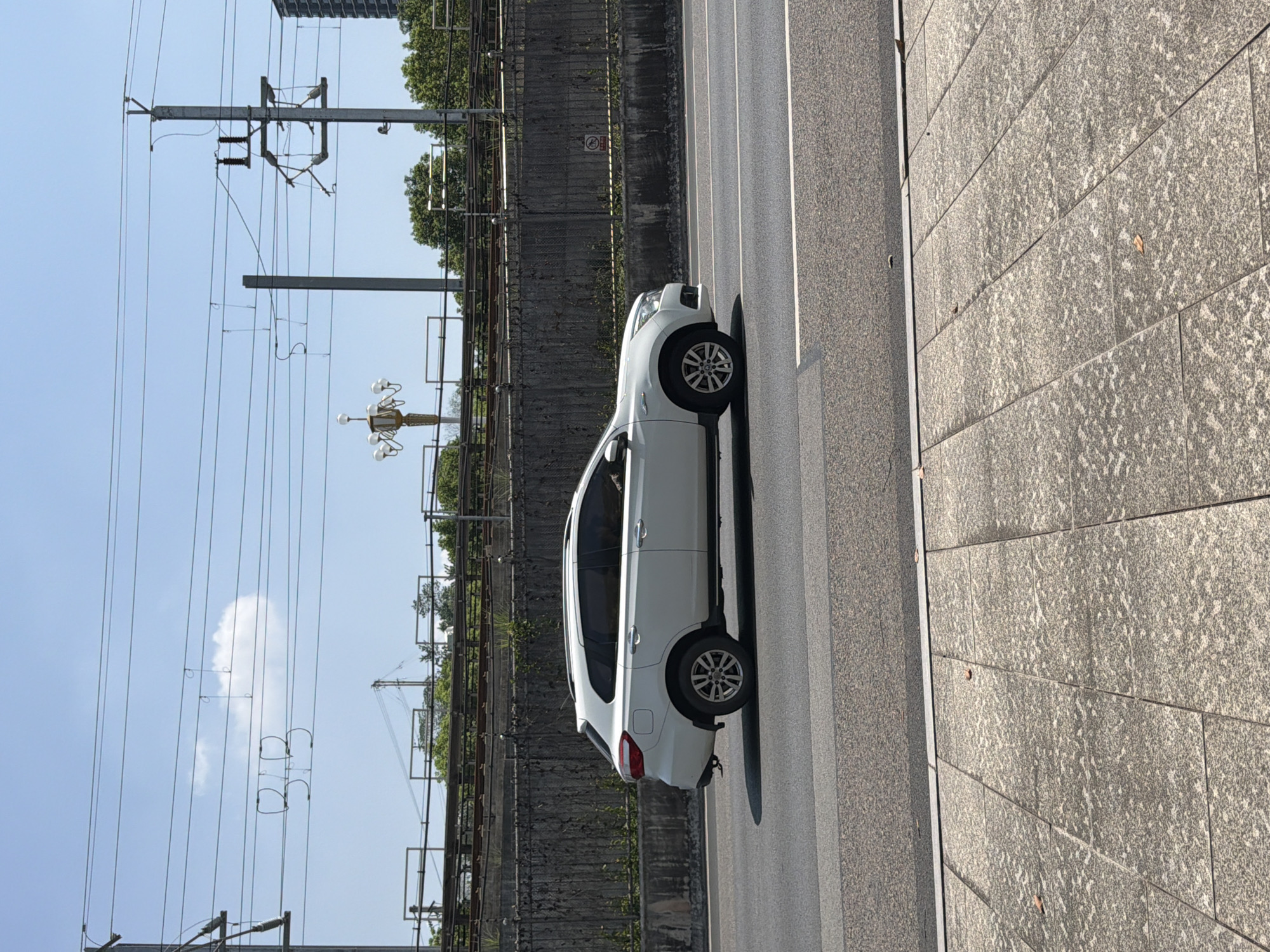 A white SUV is driving on a paved road, seen from the side. The road has multiple lanes and is bordered by a concrete barrier and a sidewalk with square, patterned tiles. In the background, there is a railway track elevated above the road, with greenery and trees beyond. Overhead, power lines crisscross the blue sky. A lamppost with multiple lights is visible in the distance. The image was taken in Jiangjin, China, under a clear sky.
