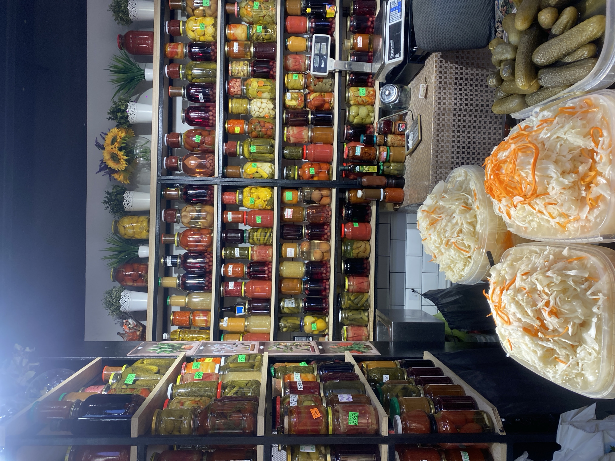 This image captures a vibrant display of preserved foods in what appears to be an indoor market or grocery store, likely in Moscow, Russia. The perspective is from slightly below, looking up at shelves and counters, with the image rotated 90 degrees clockwise.
The primary subjects are numerous glass jars filled with various preserved vegetables, fruits, and berries, neatly arranged on wooden shelves and in wooden crates. These include pickles (cucumbers, gherkins), pickled tomatoes (red and green), assorted vegetables, compotes (fruit preserves), and possibly jams or sauces. Many jars have small, often handwritten or simple printed labels, some showing numbers like "500," "700," "600," and "250" (likely prices in Rubles or weights), and words such as "АЛЕНА" (Alena, possibly a brand or producer's name) and "БОРЩЕ" (Borsch, indicating a borsch-related preserve).
On the right side of the image, large clear plastic containers hold bulk items. One container is filled with whole pickled cucumbers, while several others contain finely shredded sauerkraut with visible strips of orange carrot, ready for purchase. These bulk items are placed on a counter covered with a textured brown and gold fabric or mat. An electronic scale with a digital display and the brand name "Turan" is visible behind these containers, suggesting items are weighed for sale.
The setting features dark shelves and a dark wall on the left, contrasting with a lighter wall adorned with small potted plants and artificial sunflowers in white ceramic pots, adding a decorative touch. A section of white tiled wall is visible in the upper right background. The overall impression is one of abundance and traditional food preparation, typical of Eastern European markets. The lighting suggests an indoor environment.