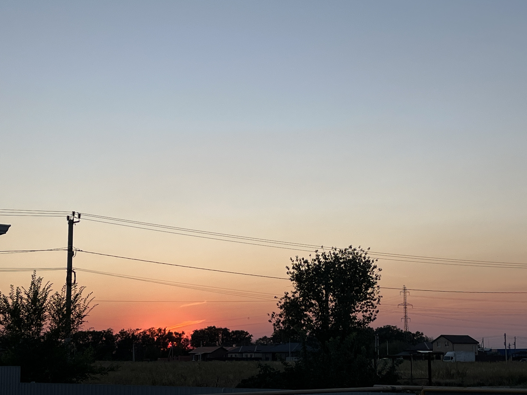 The image captures a rural or suburban sunset scene in Taganrog City District, Russia. The sky transitions from a pale blue at the top to a vibrant orange and pink gradient near the horizon, indicating the sun has recently set or is about to set. In the foreground, a dark silhouette of trees and a fence is visible. A prominent utility pole with electrical wires stretches across the middle ground. Further in the distance, the silhouettes of several houses and what appears to be a power pylon can be seen. The overall atmosphere is peaceful and serene, characteristic of twilight in a less urbanized area. No people or active events are depicted.