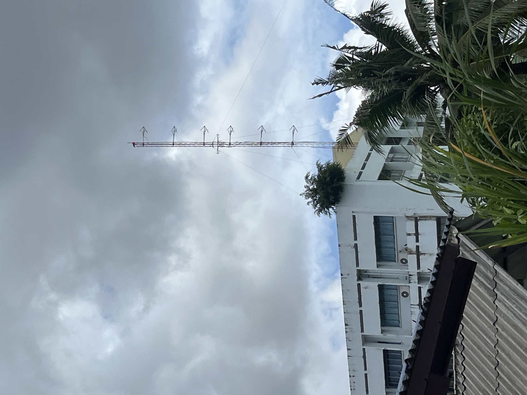 The image shows a white, multi-story building with a communication tower and a palm tree in front of it, under a cloudy sky. The building has several windows and air conditioning units visible. The communication tower is tall and slender, with multiple antennas or directional elements attached to it. Lush green foliage, including palm fronds and other plants, partially obscures the lower portion of the building and the base of the tower. The sky is overcast with a mix of grey clouds and patches of blue sky. The scene suggests an outdoor urban or semi-urban environment, possibly in a tropical location given the presence of the palm tree and dense vegetation. The general condition of the building's facade appears somewhat weathered.