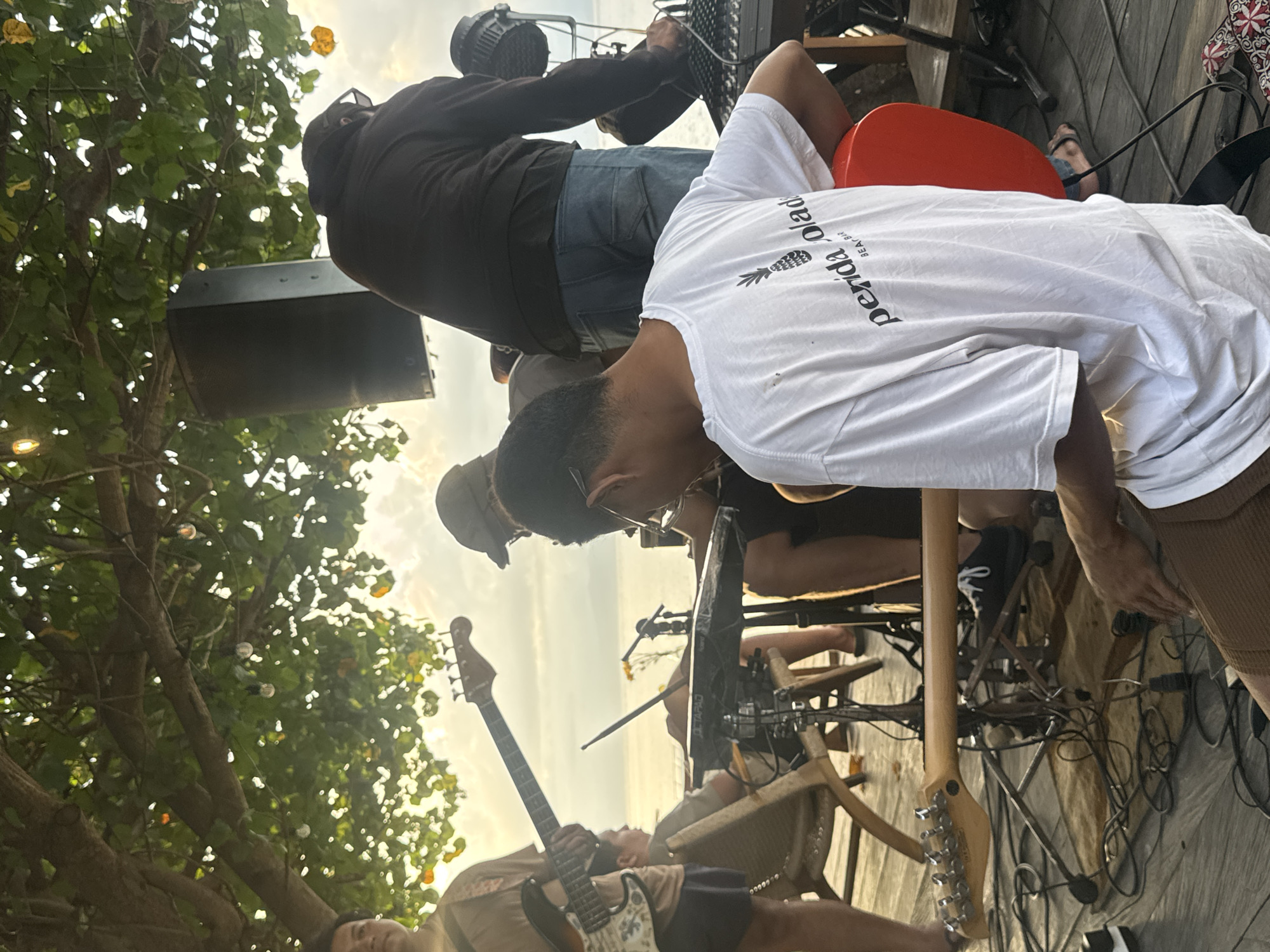 The scene depicts a band setting up or performing outdoors, likely in Nusa Penida, Indonesia, indicated by the "perida olad BEACH BAR" text on the white t-shirt of one of the individuals. A person in a brown long-sleeved shirt adjusts a stage light or equipment. Another person wearing a white t-shirt leans over, potentially adjusting equipment. The scene includes two guitarists: one playing, and one guitar resting nearby. There's a drummer behind with drum set parts clearly visible. A sound speaker is placed to the side, with tree branches and lights in the background. The ground is a wooden platform with multiple wires and music stands scattered. A red chair is also present.