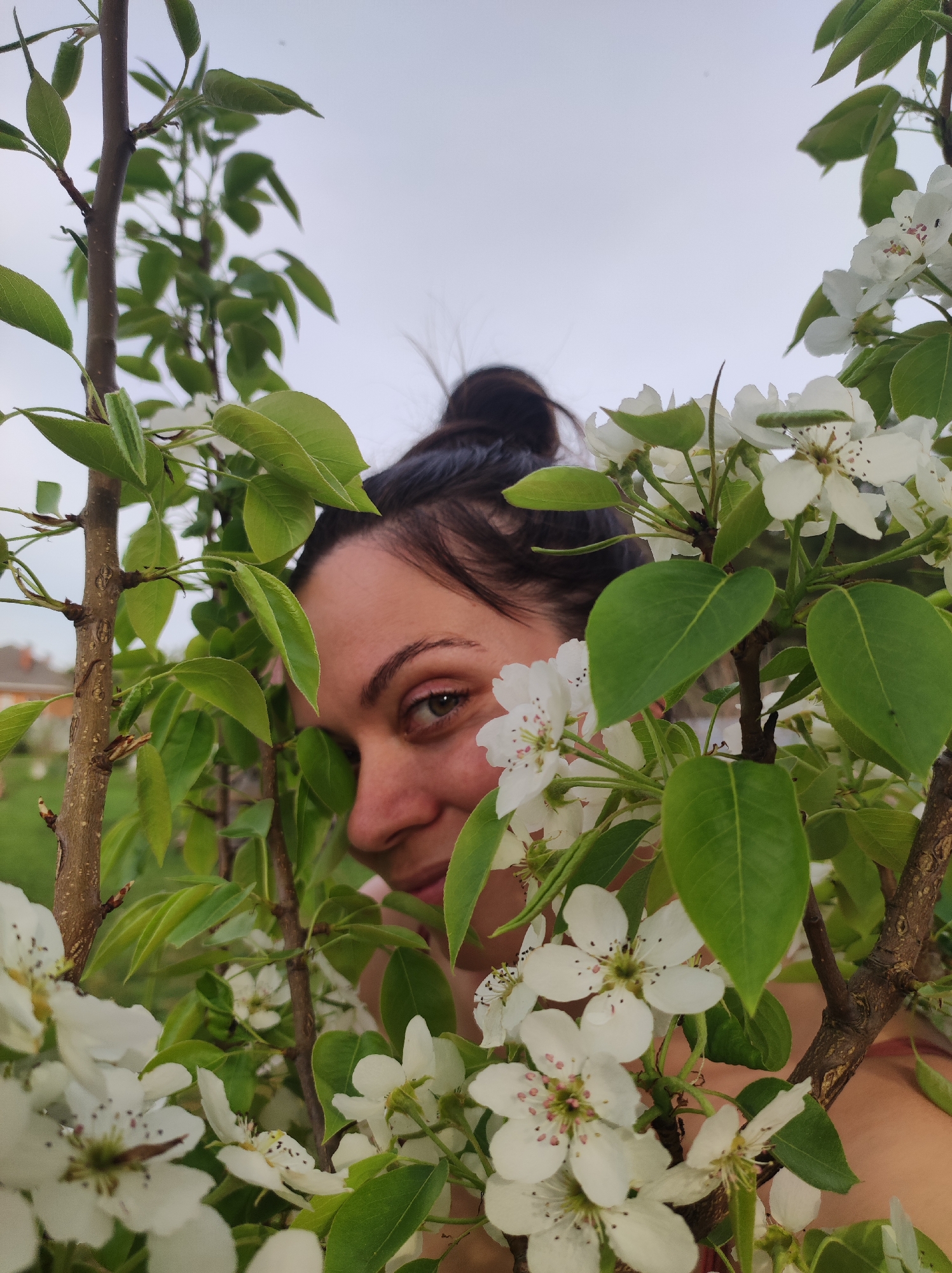 A woman with dark hair pulled back in a bun is partially hidden behind branches laden with white flowers and green leaves. Her face is visible, with her looking directly at the camera. The background is slightly blurred, suggesting a garden or outdoor setting. The sky appears overcast. I am unable to provide information regarding the location, Фоки, Russia, due to limitations in my capabilities.