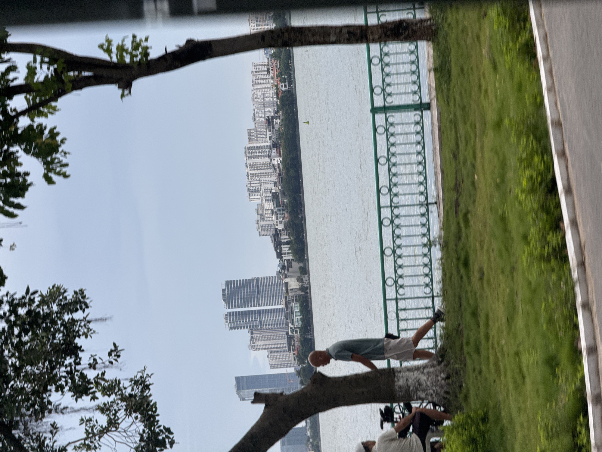 The image depicts a waterside scene in Hanoi, Vietnam, likely along West Lake (Hồ Tây), given the presence of a large body of water and modern high-rise buildings in the background. In the foreground, a man with a shaved head and wearing a light green t-shirt and beige shorts is walking along a grassy area next to a decorative green metal fence. He appears to be walking towards the right of the frame. A tree trunk with a white-banded base is to his left, and behind him, another person is partially visible, seated and wearing a white cap. Across the water, a cityscape of tall apartment buildings and other structures is visible under an overcast sky. A yellow buoy is present in the water. The overall atmosphere is calm and urban, suggesting a public park or promenade area.