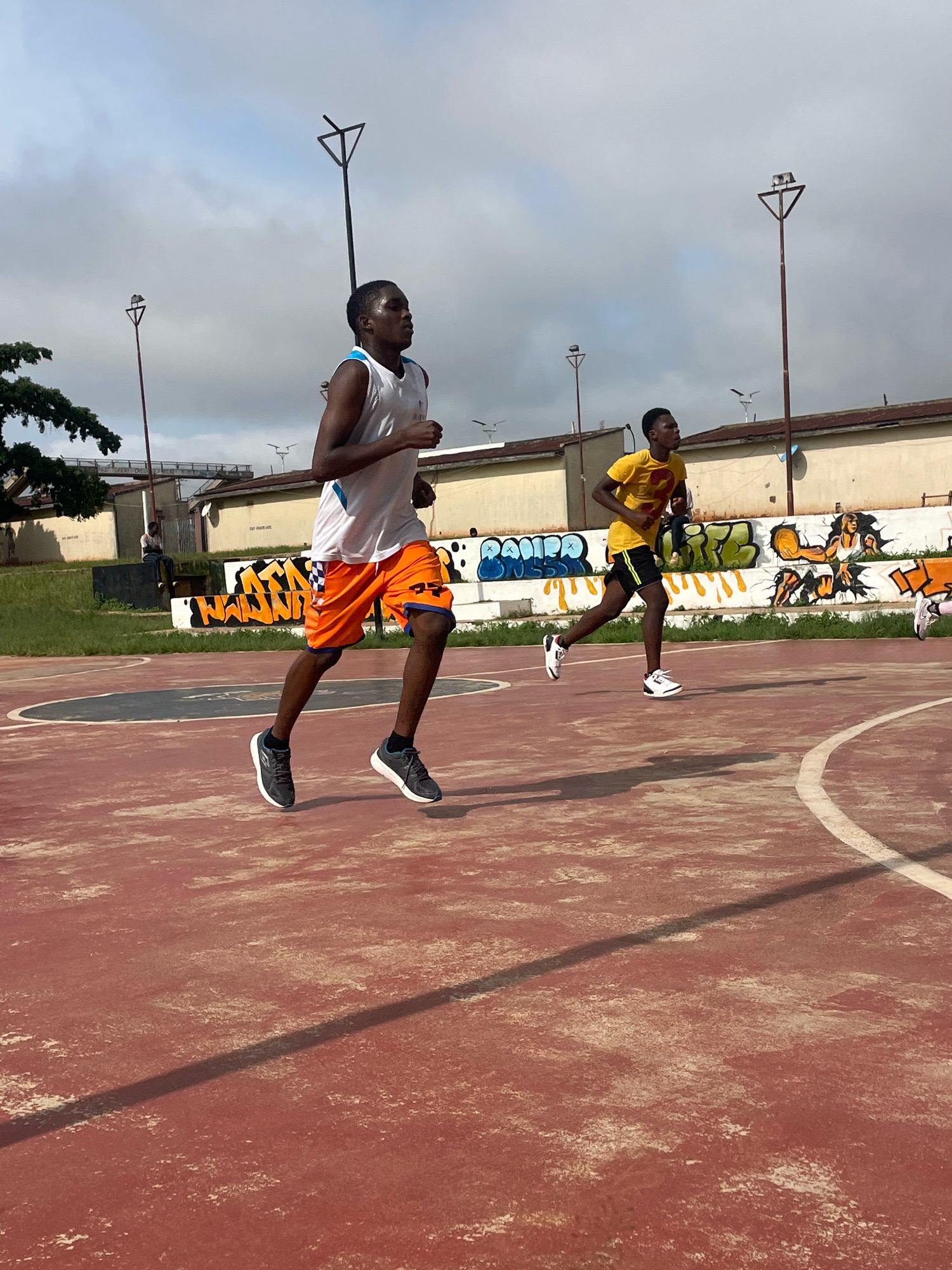 The image depicts two young men jogging on a red basketball court. The man in the foreground, on the left, wears a white tank top with blue trim and orange shorts. The man in the background, on the right, is wearing a yellow t-shirt and black shorts. Behind them, a wall is covered in graffiti. The court appears to be outdoors, with a cloudy sky overhead. Lampposts are visible in the background, and trees are to the far left. The setting is consistent with an urban environment, possibly a park or community sports facility. The image does not provide specific details to confirm the location as Gura Top, Nigeria.