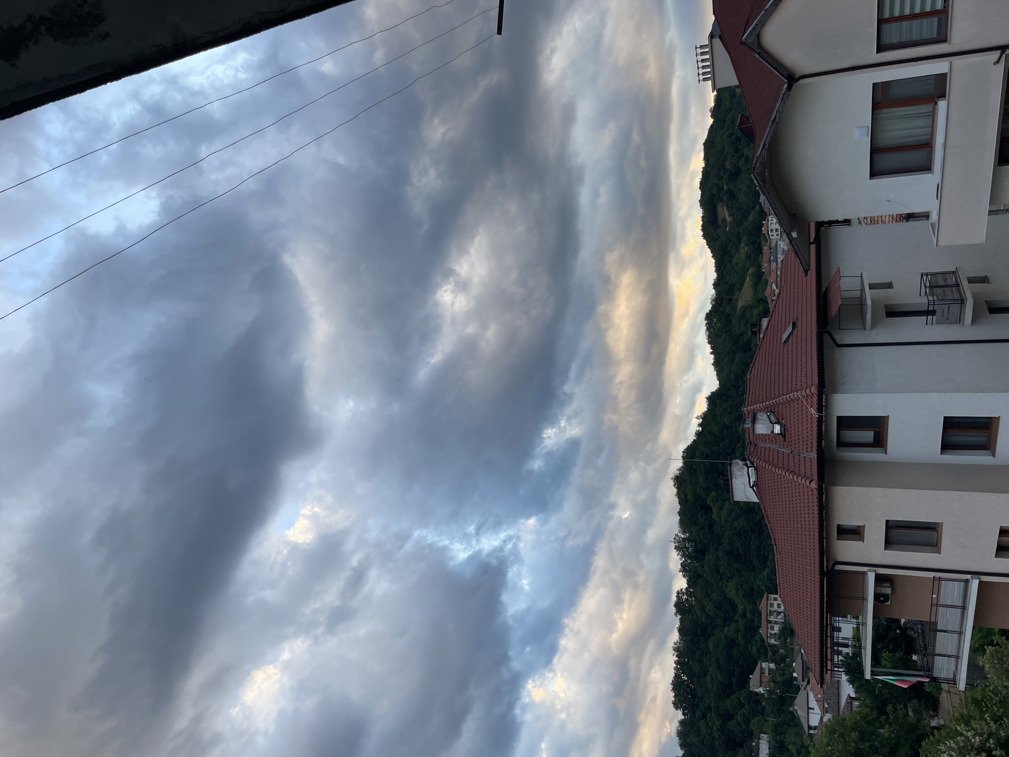 The image captures a view of buildings and a hillside under a dramatic, cloudy sky. The buildings in the foreground appear to be modern apartment complexes with red tile roofs and white or beige facades. One building features a balcony with a Bulgarian flag. In the background, a densely forested hillside is visible, dotted with houses. The sky is filled with heavy, dark gray clouds, with patches of lighter sky and a hint of yellow or orange light peeking through in the distance, suggesting the possibility of sunset or an approaching storm. Several thin, dark power lines are stretched across the upper right portion of the frame. The scene is likely from Zlatograd, Bulgaria, given the architectural style and the hilly terrain characteristic of the region. No people or animals are visible in the image.