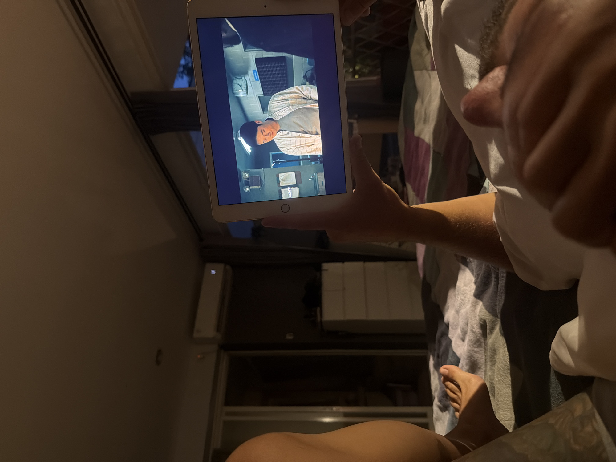 In a dimly lit bedroom in Kuta Selatan, Indonesia, an adult male lies on a bed, holding a tablet computer in his hands. The tablet displays a scene from a television show or movie, featuring an Asian man in a checkered shirt. The room has an air conditioning unit mounted on the wall and a dresser. A window in the background offers a view of the evening sky. The male's legs and feet are visible on the bed, covered by a blanket. The overall atmosphere is relaxed and intimate, suggesting a quiet evening at home.
