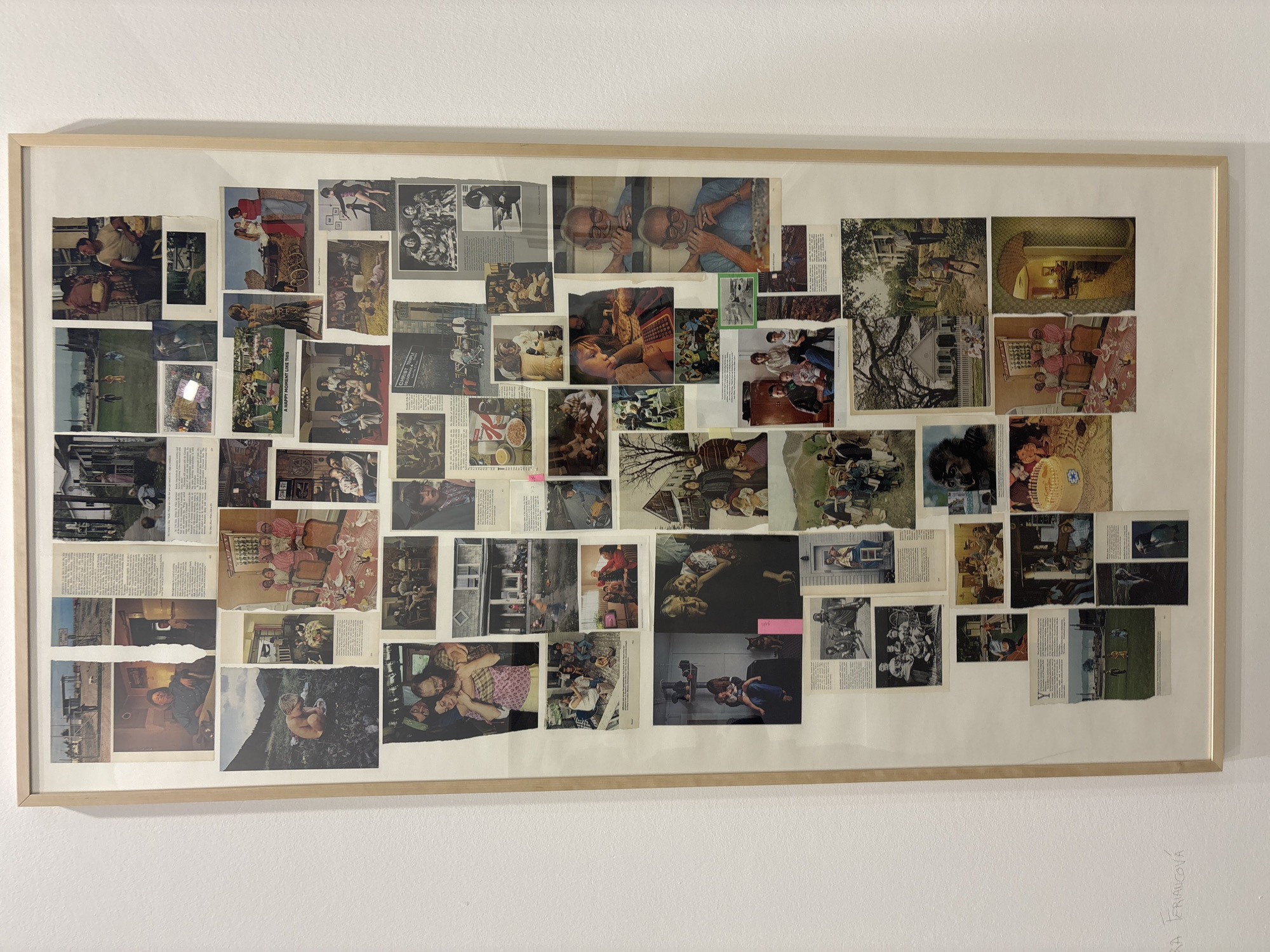 A large, rectangular framed collage is centrally displayed on a plain white wall, suggesting an exhibition setting in Milan, Italy. The frame is a light-colored wood, providing a simple border for the intricate artwork.
The collage is densely composed of numerous cut-out images and snippets of text, layered and overlapped to create a rich tapestry of scenes. The subjects predominantly feature people in various settings and activities: families gathered around tables, children playing outdoors, individuals engaged in work or leisure, and groups in both candid and posed situations. A striking central image shows two older men with glasses, mirrored, looking intently. Other elements include diverse objects such as furniture, household items, buildings, trees, and landscapes, along with some animals like horses and a primate. The images are a mix of color and black and white, often with torn edges from their original print sources.
The primary activity taking place is the display of this mixed-media art piece. Within the collage itself, a multitude of everyday life events and moments are captured, ranging from domestic scenes and social gatherings to outdoor activities.
Notable details include the collage's fragmented nature, where diverse photographic styles and eras are juxtaposed. Snippets of text are visible throughout, with one clearly legible phrase stating "A HAPPY MOMENT LIKE THIS" and another appearing as a title or heading, "GHOSTLY TRIPLE". The artwork creates a sense of collective memory or narrative through its assembled fragments. On the white wall, outside and to the lower right of the frame, faint handwritten text in pencil is visible, which appears to be the artist's name: "SARA FRIACÓVA".