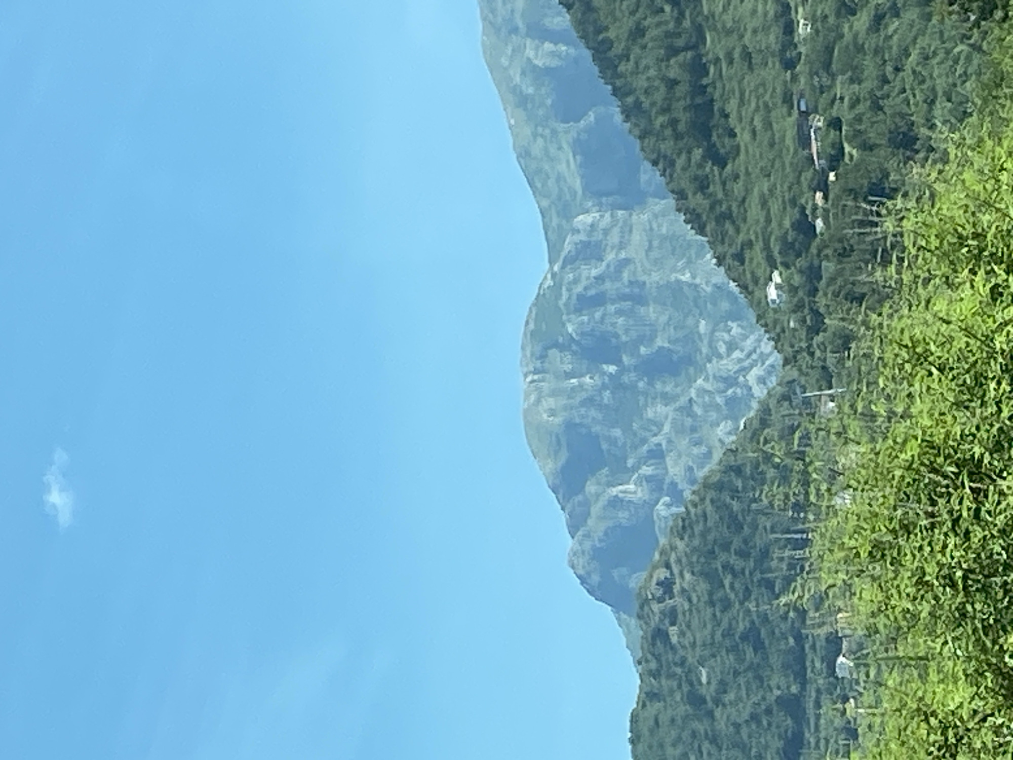 The image depicts a scenic mountain landscape under a clear blue sky with a single small cloud. In the foreground, lush green foliage dominates the lower portion of the frame. Beyond the foliage, rolling hills covered in dense green trees rise, dotted with a few small white buildings, suggesting a rural or semi-rural settlement. The mid-ground features a prominent, rugged mountain with distinct rock formations and steep cliffs. The background is composed of layers of receding mountain peaks, also covered in greenery, fading into the distance. The overall scene suggests a natural, mountainous environment, likely in or near the city of Dub, Montenegro, known for its mountainous terrain and proximity to natural beauty.