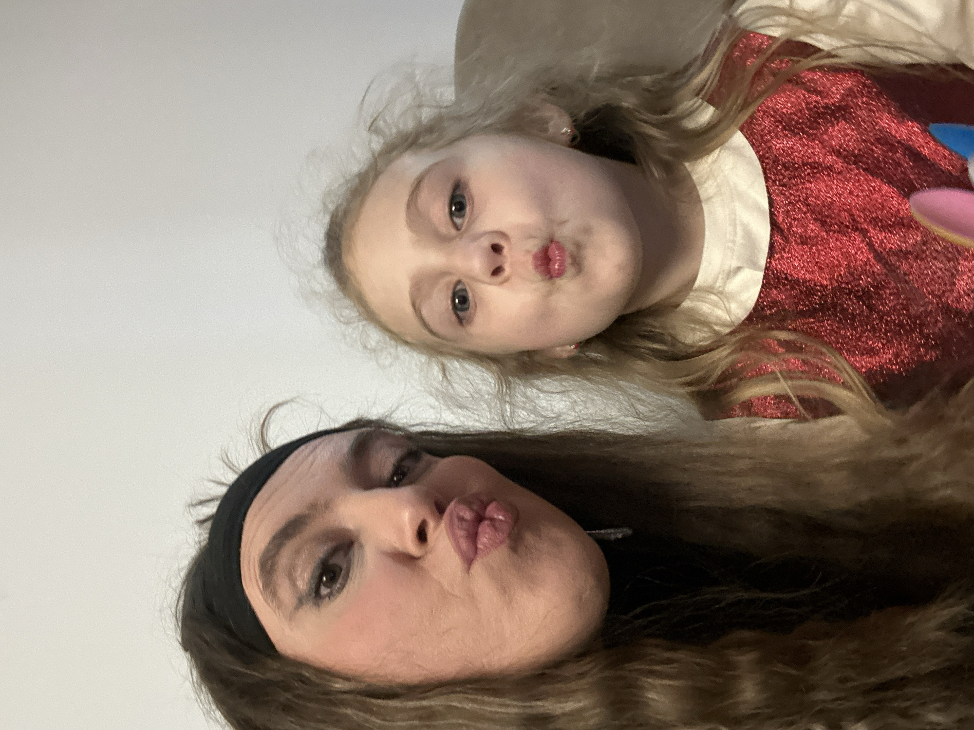 Two people are in the image: a young fair-skinned child with blonde hair and an adult fair-skinned woman with brown hair and a black headband. Both are looking at the camera, and making a kissing face with their lips. The child is wearing a red glittery top with a white collar. The background is a white wall. The location is indeterminable.