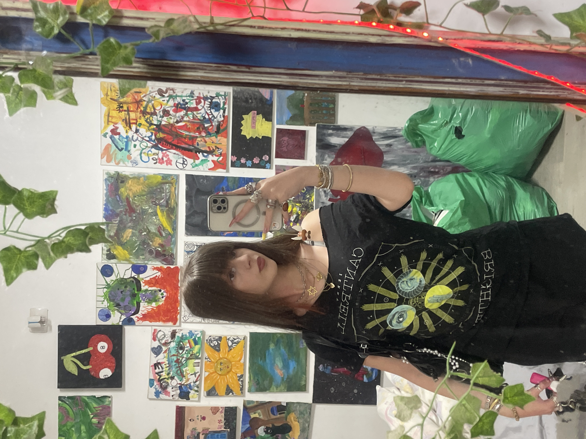 A person with shoulder-length dark hair is taking a mirror selfie in a room with a collection of small paintings or artwork mounted on the wall. The artwork varies in style and subject matter. The person is wearing a black t-shirt with a yellow graphic design. They are holding a smartphone in their left hand, and their arm is adorned with bracelets and rings.
There are artificial vines and red string lights framing the mirror. In the upper-right corner of the reflection are green plastic bags.
There is no information in the image to indicate the location is in Raanana, Israel.