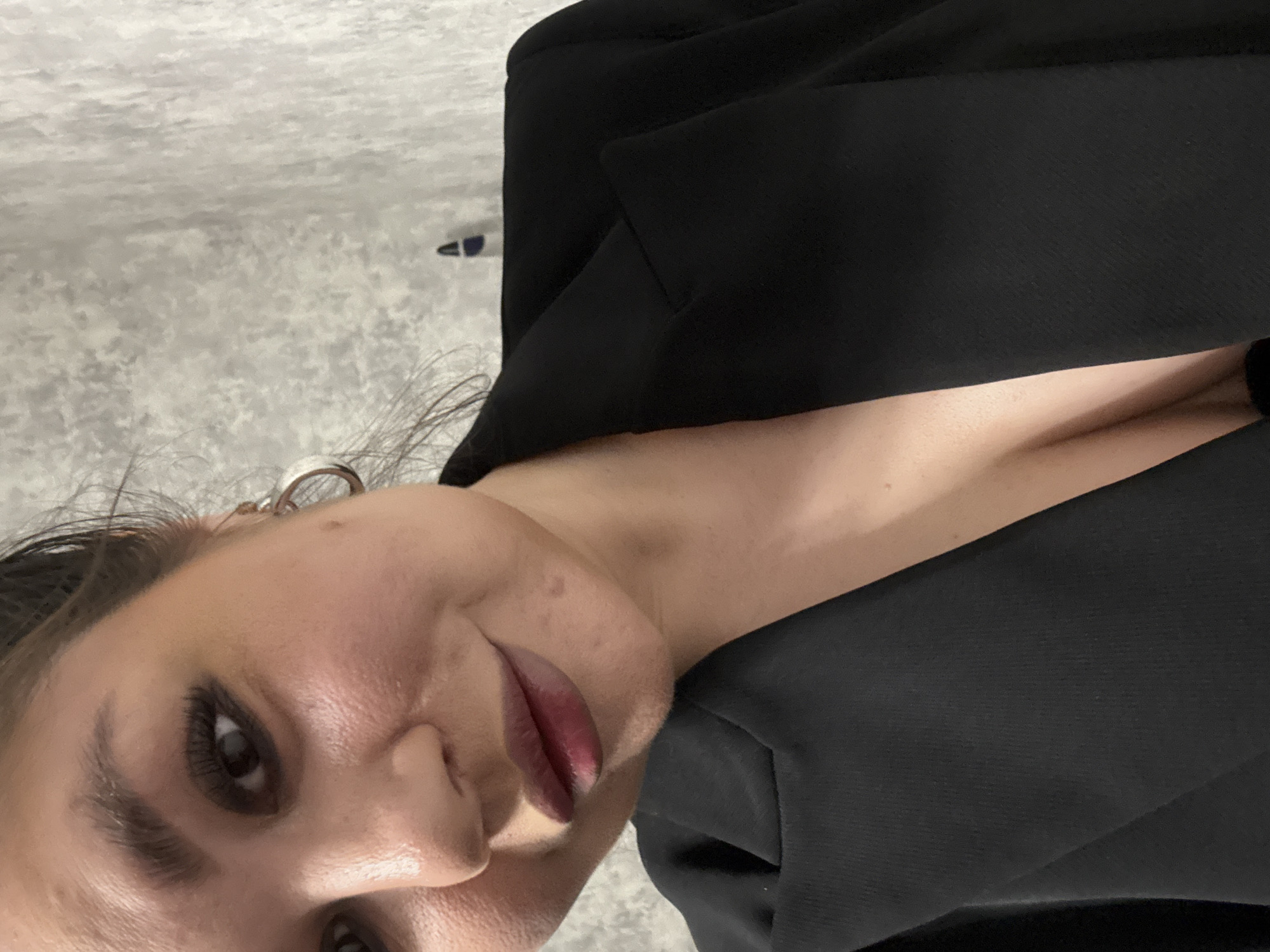 A close-up, rotated view of a woman with fair skin, dark eyes, and full lips. She is wearing a black top, possibly a jacket, with a low-cut neckline. An earring is visible on her left ear. The background appears to be a textured surface. The location is not discernible from the image.