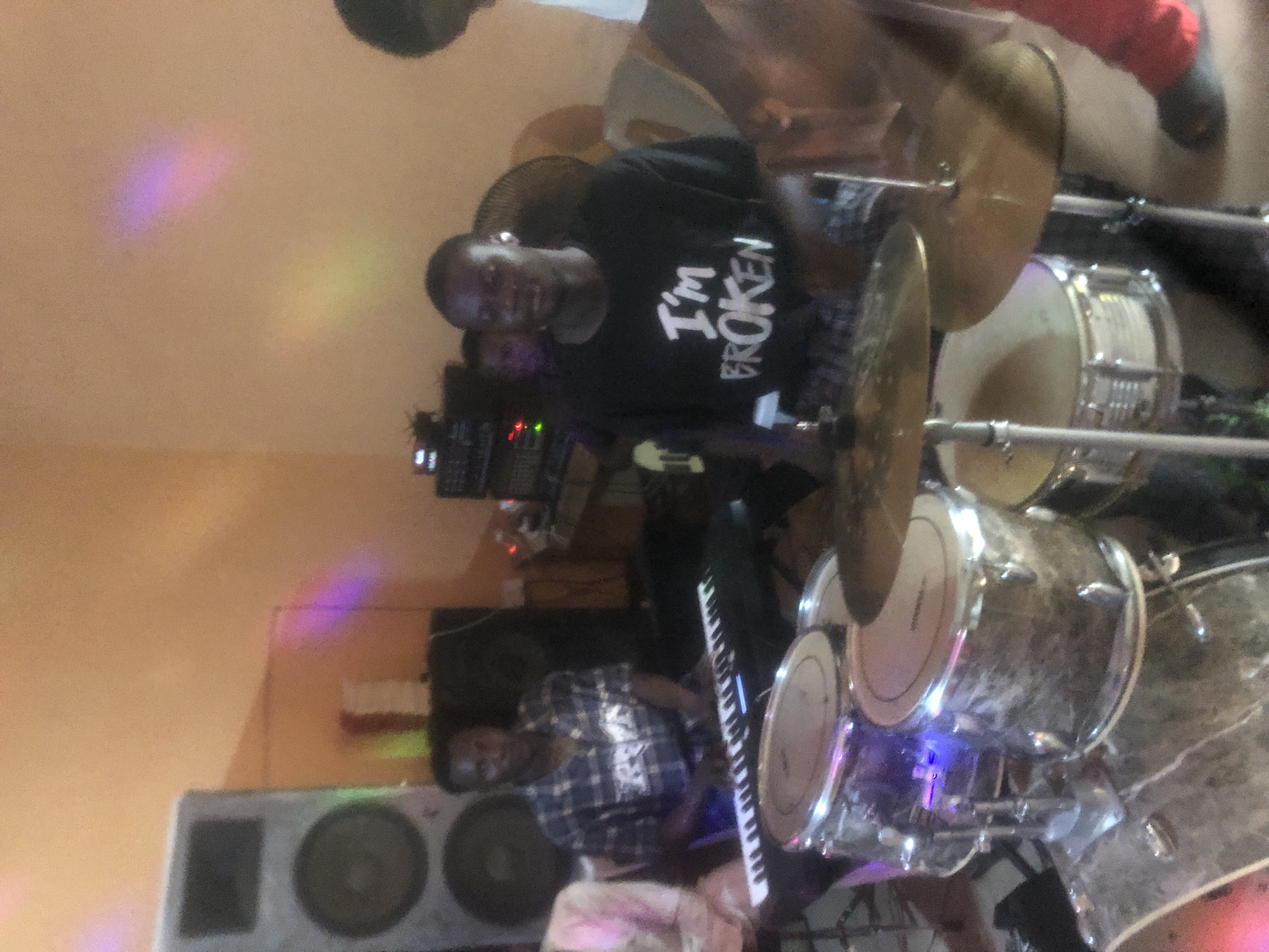 The image shows a band performing indoors, likely in Nkwelle Ezunaka, Nigeria. A drummer in a black t-shirt with the words "I'M BROKEN" is positioned behind his kit. A keyboard player in a plaid shirt is visible next to the drums. Speakers and other music equipment are also present. Multicolored light patterns illuminate the ceiling.