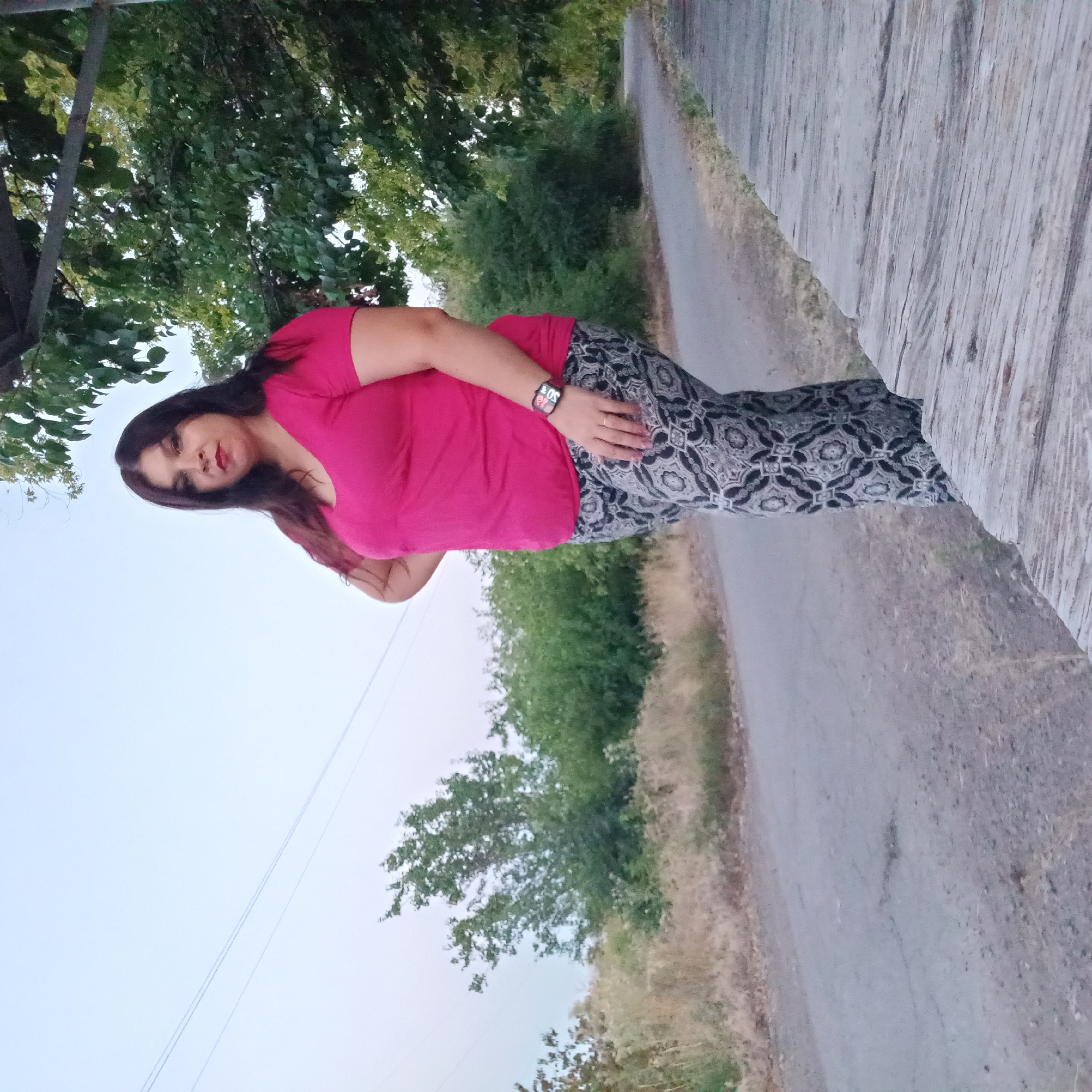 In the city of Berezhynka, Ukraine, a woman stands by a paved road, with trees and foliage surrounding the area. The woman, with dark hair and red lipstick, wears a pink t-shirt and patterned pants. She has one hand on her hip and a watch on her wrist. The road appears to be a country road, with dry grass and bushes lining its sides. The sky is overcast, suggesting it might be evening or a cloudy day. A wooden structure is visible to the right of the woman, providing some shade. The overall scene depicts an outdoor setting with a natural ambiance.