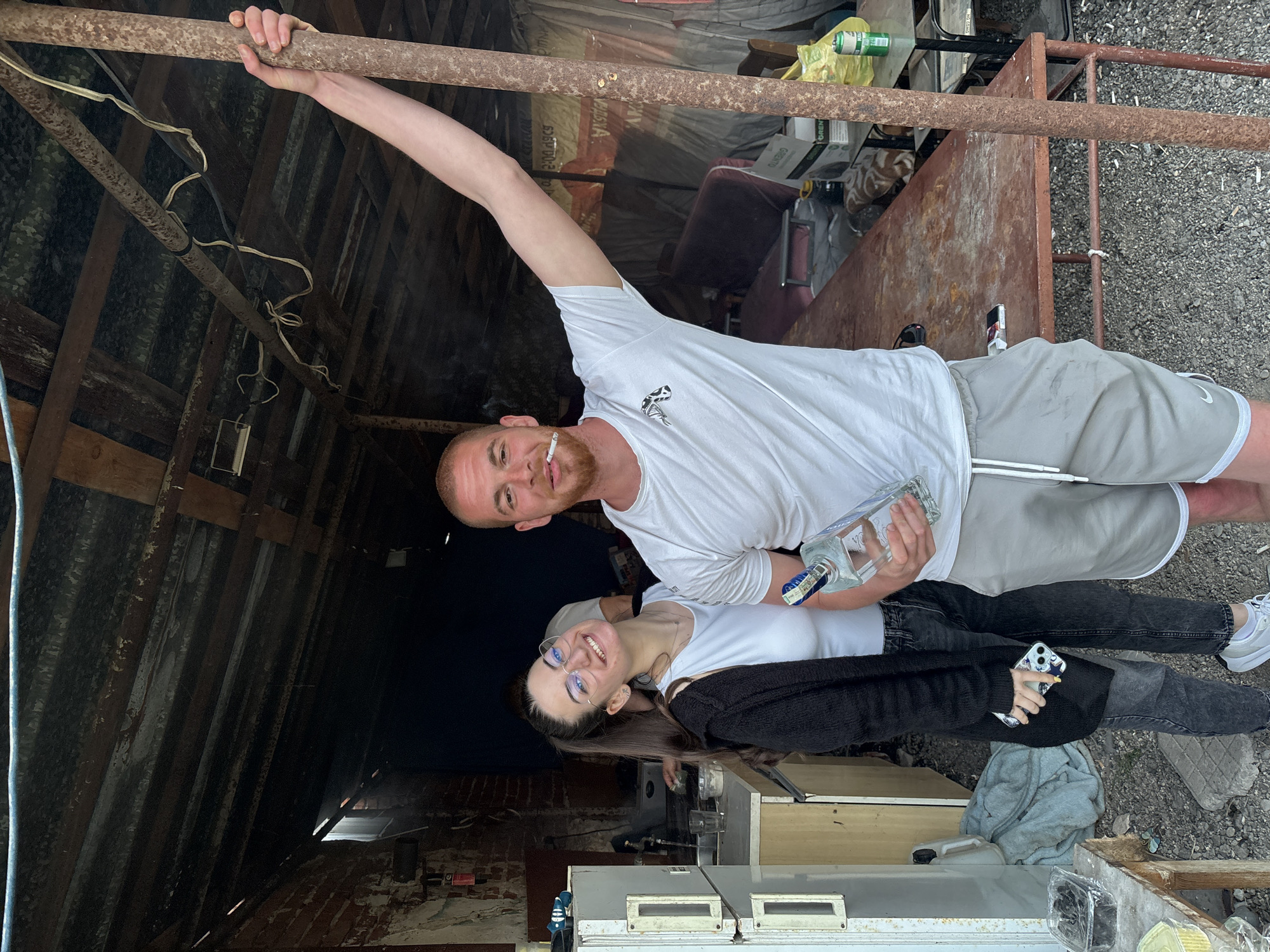 The image shows two people standing outside in an area containing what appears to be scrap metal and old building materials. A man with a cigarette in his mouth is holding onto a metal bar, which appears to be part of a structure. He is wearing a white t-shirt and gray shorts, and he is also holding a bottle of alcohol. A woman with glasses is standing next to him, wearing a white t-shirt, dark jeans, and a dark sweater. She is holding a smartphone. In the background, there is a dilapidated structure with a corrugated metal roof. The environment includes metal beams, debris, and what seems to be an outdoor area with scattered objects. There are also items like a refrigerator and containers, suggesting the location is in an urban environment and not a designated business. There is no indication in this photo where the subjects are located or indications of the city Chirpan, Bulgaria.