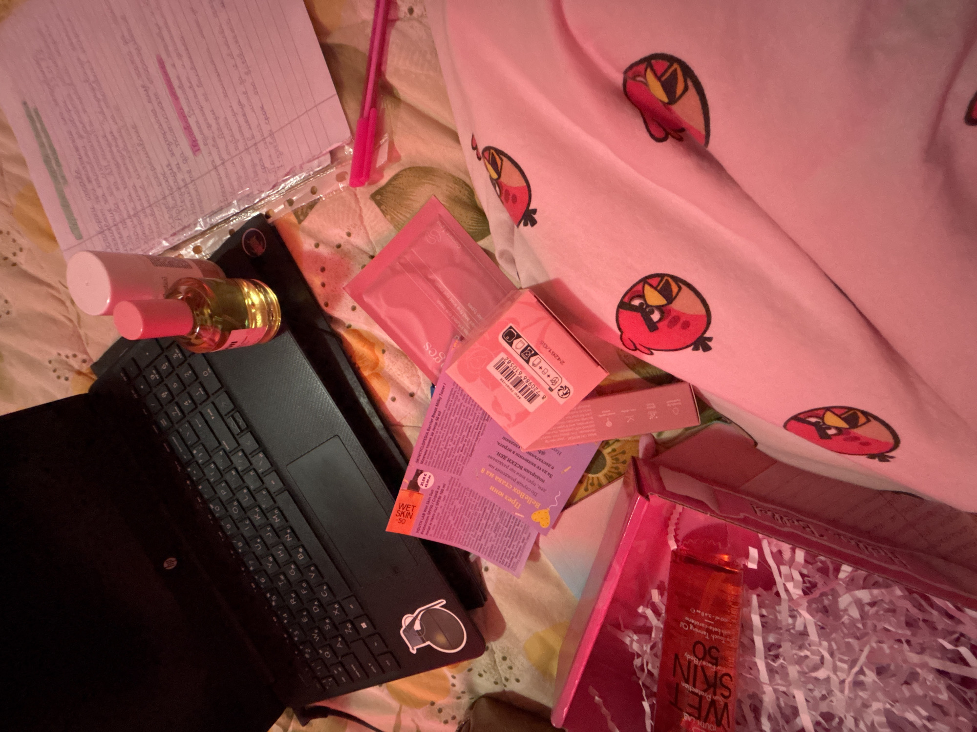 The image depicts a cluttered scene on what appears to be a bed or similar soft surface. There is a laptop, a bottle of tanning oil and moisturizer, skincare products in boxes and packets, handwritten notes, and a pen. The environment is characterized by a warm, pinkish lighting. The presence of Cyrillic script on several of the product labels suggests the scene is located in a country that uses the Cyrillic alphabet, such as Bulgaria. However, the specific location within Garmen, Bulgaria cannot be determined from the provided image.