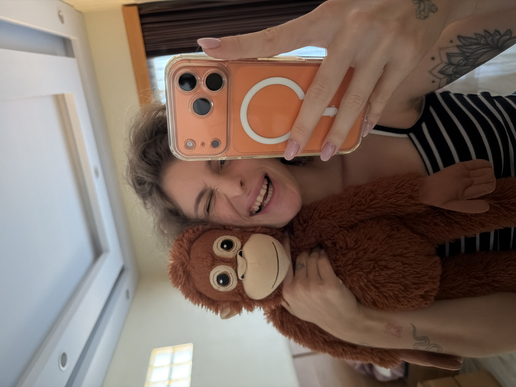 A young woman with curly brown hair is taking a selfie in a room. She is holding a brown stuffed monkey in her left arm and her right hand is holding an orange phone. She is winking and smiling at the camera. She is wearing a black and white striped tank top. Her left arm has a snake tattoo and a red stamp tattoo. Her right arm has a floral tattoo. The room has a white ceiling with recessed lighting. A window with a frosted glass pane is visible on the left side of the frame. A brown curtain is visible in the background on the right. The scene appears to be an indoor residential setting in North Kuta, Indonesia.