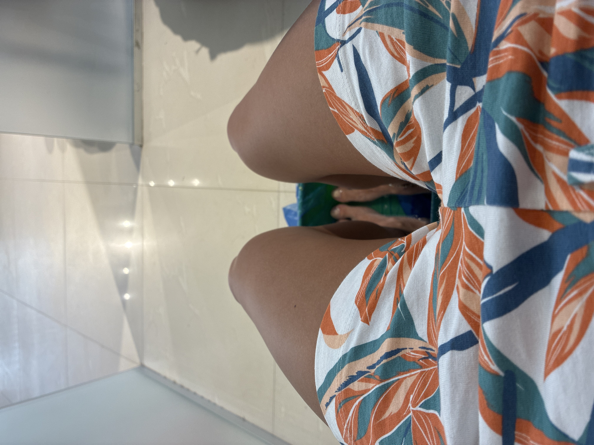 A first-person perspective shot shows a person's legs, clad in white shorts with an orange, green, and blue leaf pattern, resting in what appears to be a nail salon. The person's feet are submerged in a green foot bath, visible just below the hem of the shorts. The floor is tiled in a light, neutral color, and the overall environment suggests a clean, well-lit indoor space, possibly in Khashuri, Georgia, a city known for its diverse landscape and growing tourist industry.