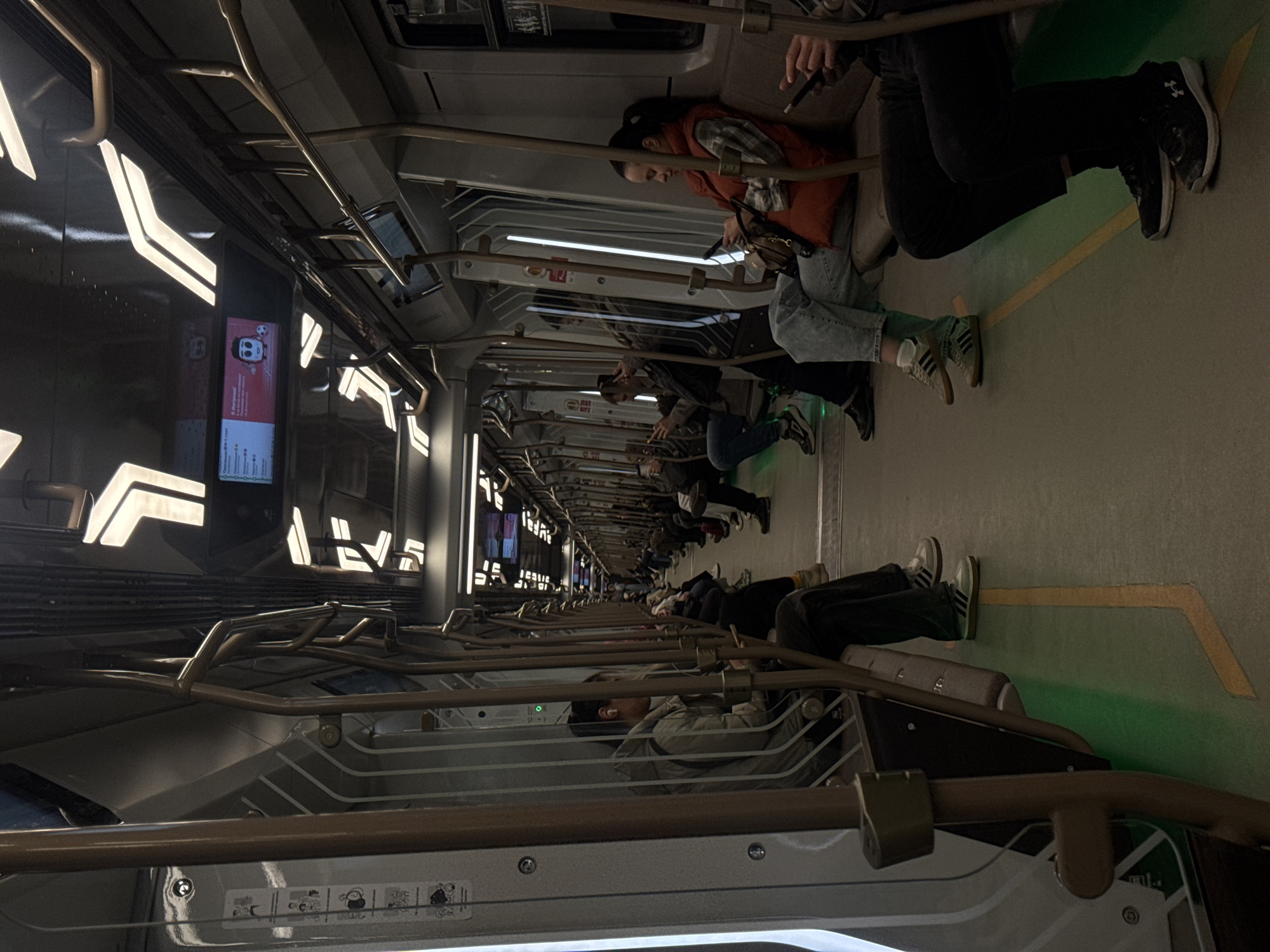 The image shows the interior of a Moscow Metro train car, Russia. Passengers are seated along the sides of the train, visible in a line along the right side of the frame. Many are looking at their phones. The car is modern, with gray and black walls, illuminated patterns along the top, and overhead handrails. The floor is a greenish gray color with yellow markings. Displays on the left side of the train provide information, including routes, and safety guidelines.