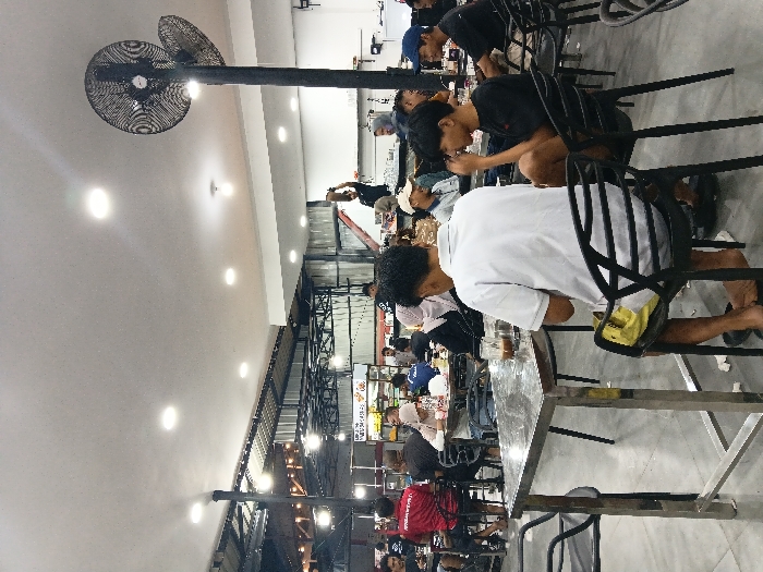 The image depicts an indoor food court or large casual dining area located in the city of Seumadam, Indonesia. The floor is light grey, and the ceiling transitions from a flat white surface with embedded spotlights in the foreground to an exposed industrial-style corrugated metal roof with suspended lighting in the background.
Approximately twenty-five individuals are visible, primarily adult males, seated at various tables. In the immediate foreground, two males occupy dark plastic chairs at a rectangular metal table with a reflective surface. The male with his back to the camera wears a white short-sleeved polo shirt and yellow shorts; a glass containing a dark beverage is on the table in front of him. The male seated to his right wears a black short-sleeved t-shirt and dark shorts, with his hand near his mouth. Other patrons include individuals in red and black t-shirts and a female wearing a light-colored head covering (hijab).
In the mid-ground, a person in a dark shirt is standing on a red and white step ladder or platform, holding an object above their head, possibly engaged in maintenance or setup. Two large, black, industrial-style oscillating fans are mounted on a tall black pole positioned on the right side of the frame, extending towards the ceiling. Multiple food stalls with partial Indonesian signage and various equipment are visible in the background. The scene suggests a busy social and dining environment.