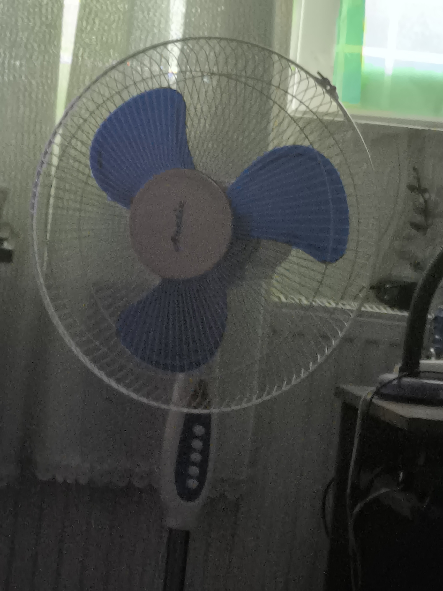The image displays a pedestal fan with blue blades and a white base, positioned indoors. The fan is the central subject, with its metallic grill and spinning blades visible. Behind the fan, sheer curtains and a window with some light filtering through suggest a room setting. To the right of the fan, a dark-colored piece of furniture, possibly a table or cabinet, is partially visible, with a black cable running across it, indicating an electrical appliance nearby. The overall lighting is dim, and the image has a grainy texture. The location is identified as Dobrogea Veche, Moldova. There are no people or specific actions depicted, focusing solely on the static scene of the fan and its surroundings.