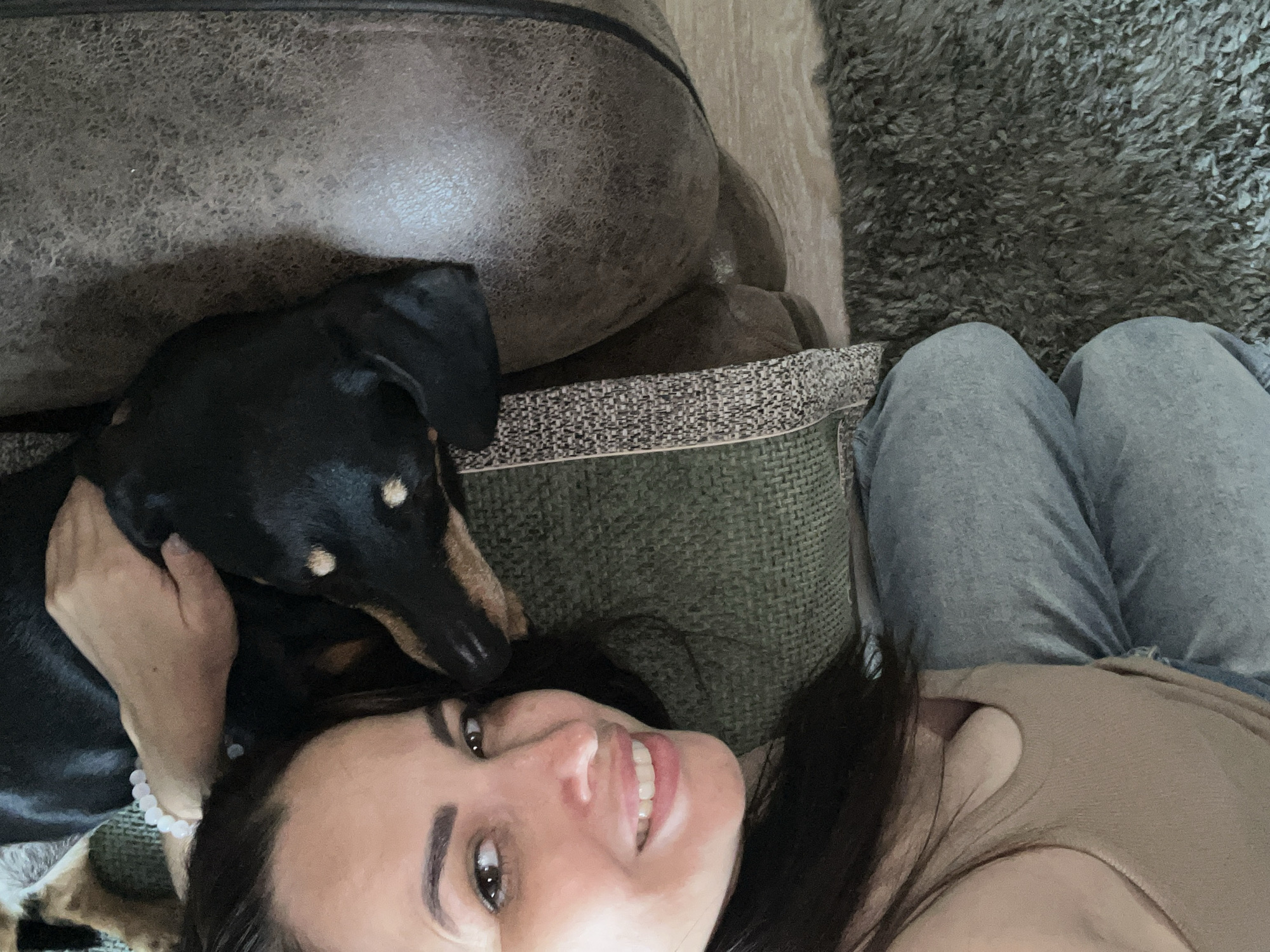 A woman and a black and tan dachshund are lying on a sofa and rug. The woman is in the foreground, smiling at the camera. The dog is resting its head on her arm. The sofa is a dark brown leather, and the rug is a dark grey shag. The scene is lit by natural light. The woman is wearing a tan tank top and grey sweatpants.
