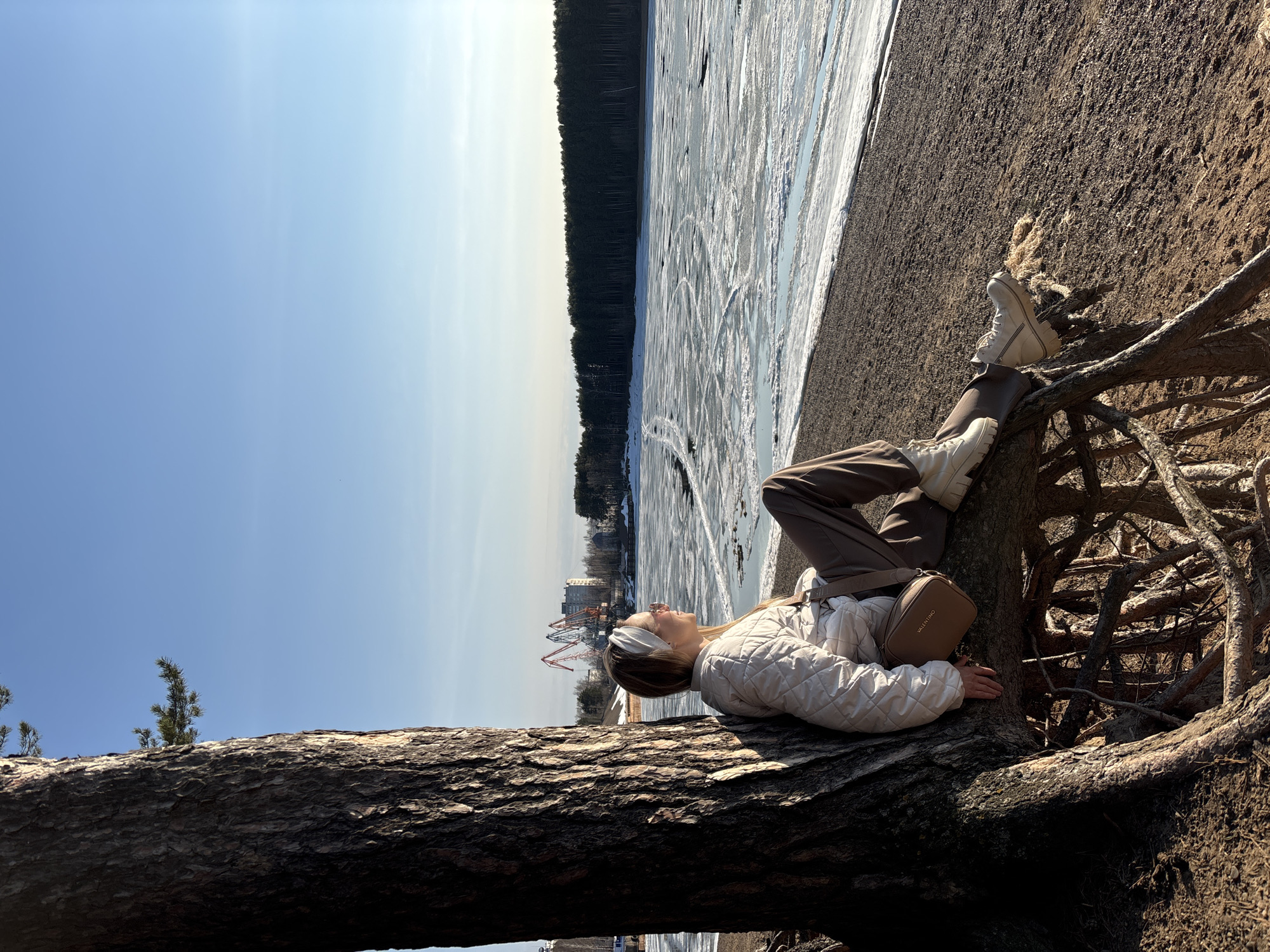 A woman rests on a fallen tree trunk along the shoreline of a frozen body of water. The woman is wearing brown pants, a white quilted jacket, and white boots. She has a light brown purse slung across her body. The background shows a landscape with a frozen lake or river covered in tire tracks, a line of trees, and some buildings, including cranes that suggest a port or industrial area in the distance. The sky is blue with some light cloud cover. The photograph depicts a scene in the city of Chaikovsky, Russia, where the body of water is known as the Kama River.