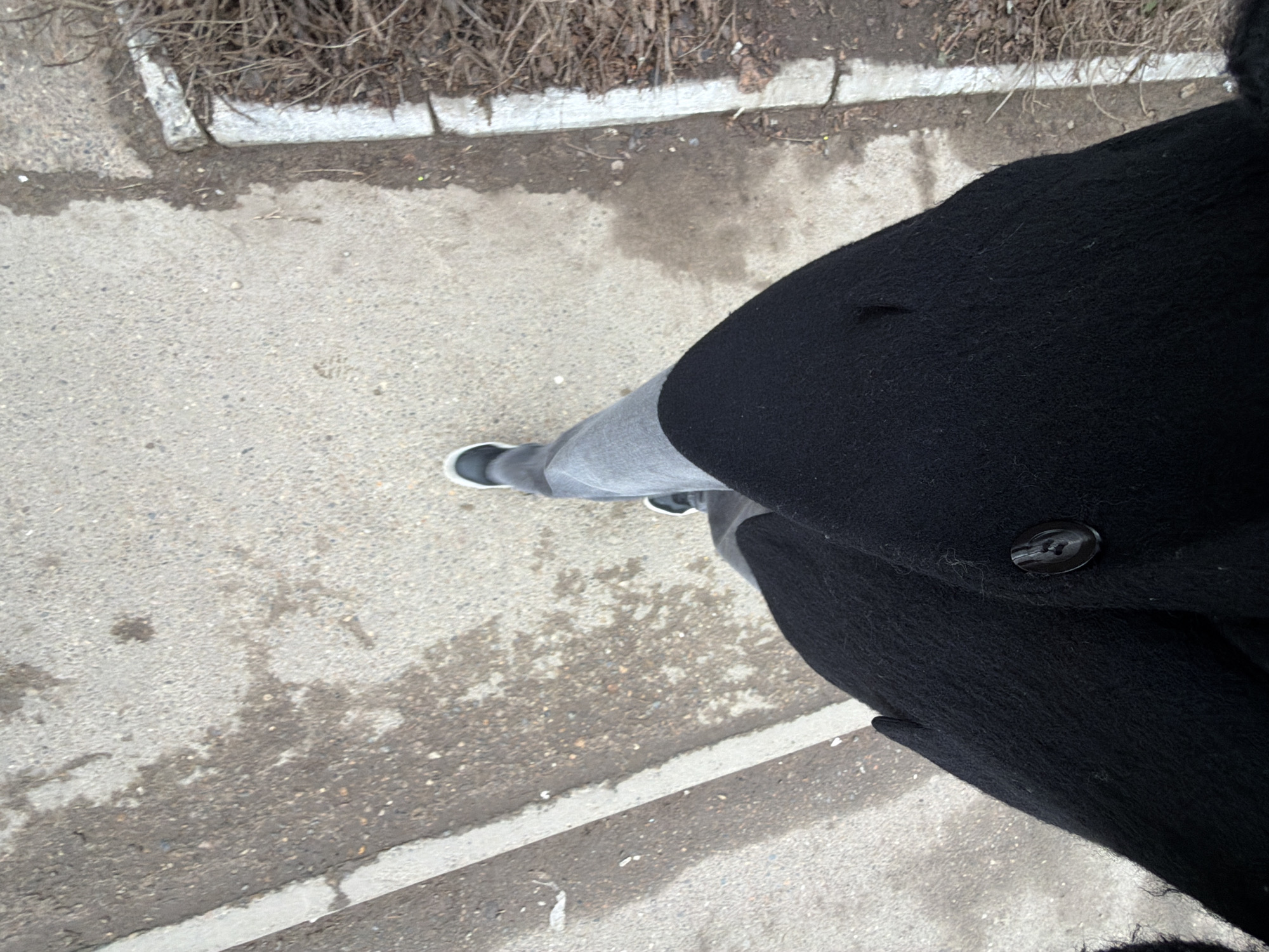 A high-angle photograph captures a person standing on a concrete sidewalk next to a curb. The sidewalk has visible water stains and a curb with small bushes. The person is wearing grey jeans, black and white shoes, and a long black wool coat. The scene could possibly be in Chaikovsky, Russia, although there is no identifying information to confirm the exact location.