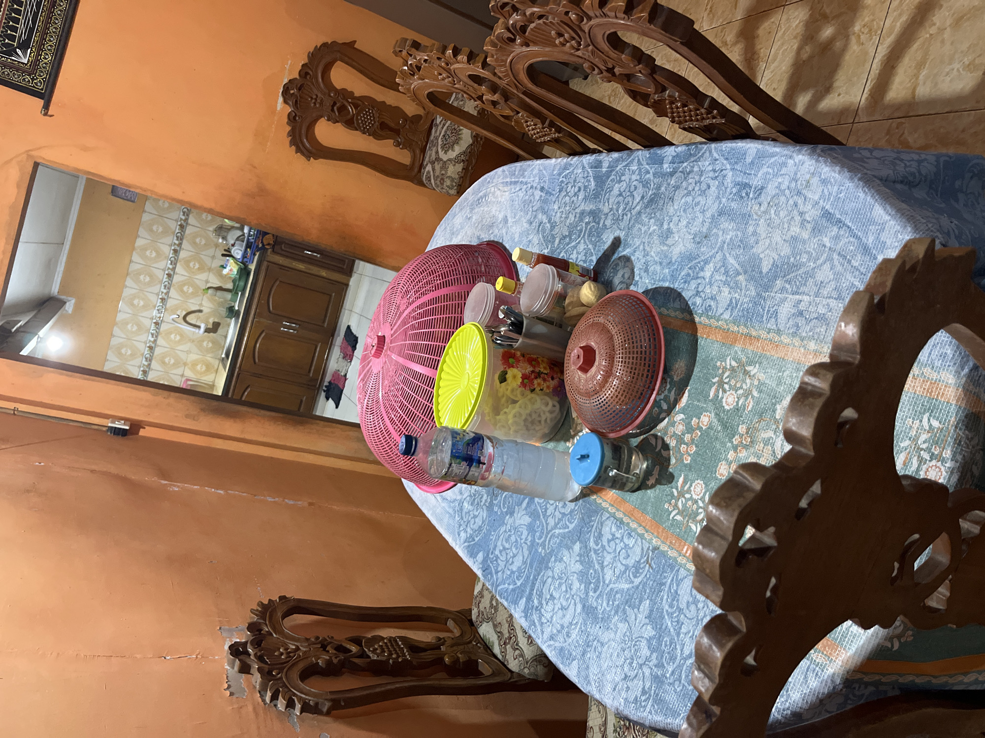 The image shows a dining table covered with a blue tablecloth patterned with white floral designs. On the table are several items, including a large pink basket-like food cover, a bright yellow lidded container filled with snacks, a bottle of water, a small glass with a blue lid and a clear liquid, a red basket-like food cover, and a few small jars and containers, possibly for spices or condiments.
The table is surrounded by several wooden chairs with intricate carvings. Two chairs are clearly visible in the foreground, and parts of two more are visible on the right side of the image. The chairs have ornate backs and seats covered in a patterned fabric.
The background reveals an open doorway leading to a kitchen area. The kitchen has wooden cabinets, a sink with a faucet, and patterned tile backsplash. The walls in the dining area are painted orange and appear to have some imperfections and peeling paint.
The lighting suggests an indoor setting, possibly during the day or with artificial lighting. The overall scene depicts a dining area in what appears to be a residential home. Given the context, this scene is likely located in Glagah Kulon, Indonesia, a region known for its residential areas and traditional homes.