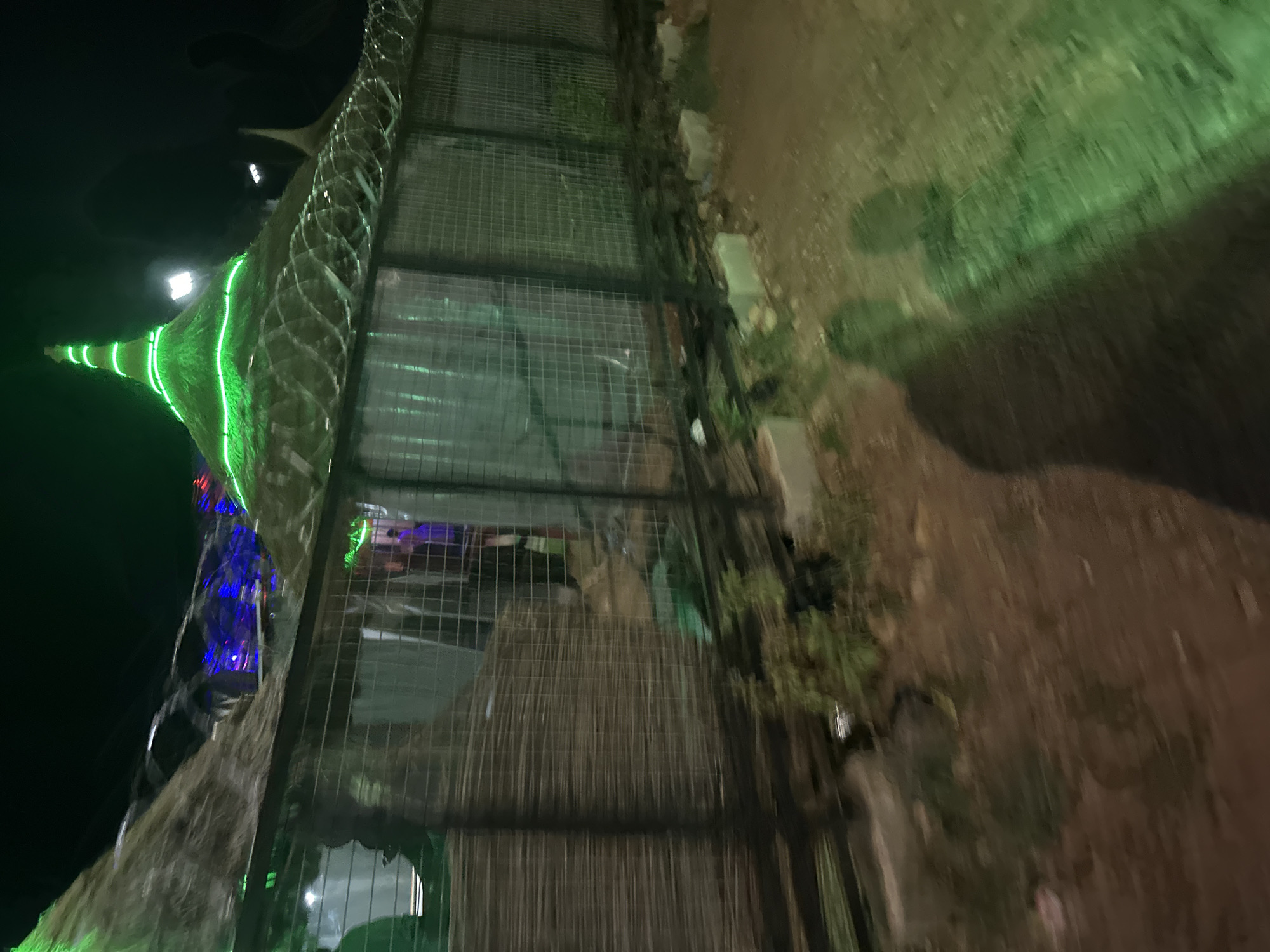 At night in Jos, Nigeria, a green-lit tower-like structure is enclosed by a wire fence. The structure, also decorated with blue lights, appears to be adjacent to a concrete wall covered in vegetation. The ground is dirt.
