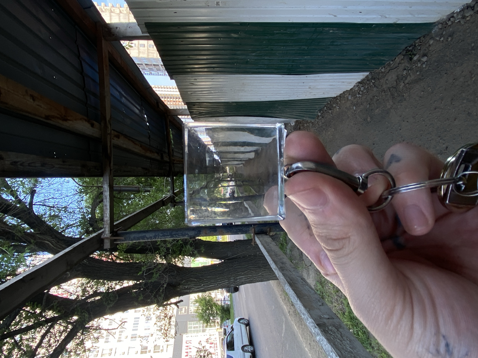 The image shows a close-up of a hand holding a glass cube, which is acting as a lens to view a scene. The scene viewed through the cube is a reflection of an alleyway with trees, buildings, and a road with a white car. The hand is wearing a silver ring and has a small tattoo on the finger. The surrounding environment appears to be outdoors, with a corrugated metal fence and some wooden structures visible. The time of day seems to be daytime, possibly late morning or early afternoon, given the bright sunlight. The weather appears to be clear and sunny. The overall impression is one of an urban or semi-urban environment, possibly in Khabarovsk, Russia, based on the provided context. The image's orientation is rotated 90 degrees clockwise.