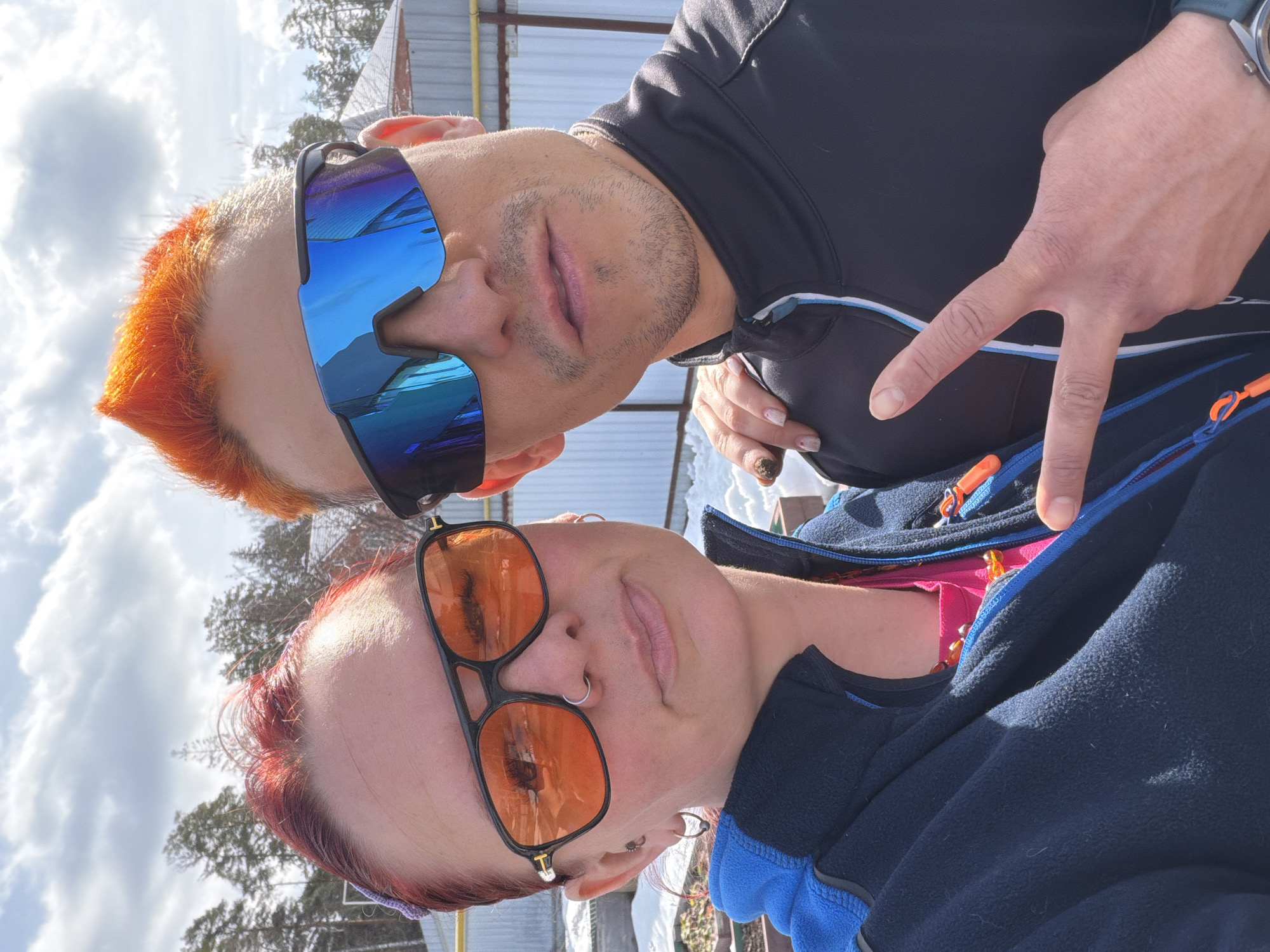 Two individuals are posing for a selfie, seemingly outdoors on a sunny day. The person at the top of the frame has short, bright orange hair and is wearing reflective blue sunglasses. They are wearing a black zip-up top and making a "peace" sign with their fingers. The person at the bottom has red hair and is wearing orange-tinted sunglasses. They are wearing a dark blue jacket over a pink garment.
The background includes bare trees, snow on the ground, and a metal structure. Based on the environment and the description, this image might have been taken near a park or recreational area in Chaikovsky, Russia during a winter or early spring season. Further details would require more visual information.