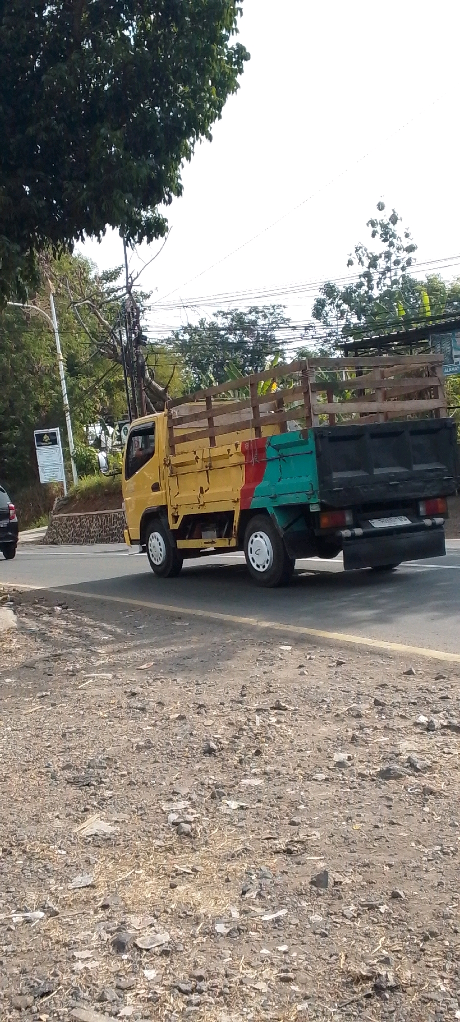 A yellow, blue, and red truck is seen driving on a road in Cilaku, Indonesia. The truck has a wooden stake bed and appears to be carrying cargo. The road is paved and there are trees and power lines visible in the background. The ground beside the road is covered in gravel and dirt. There is a white signpost on the left side of the road.