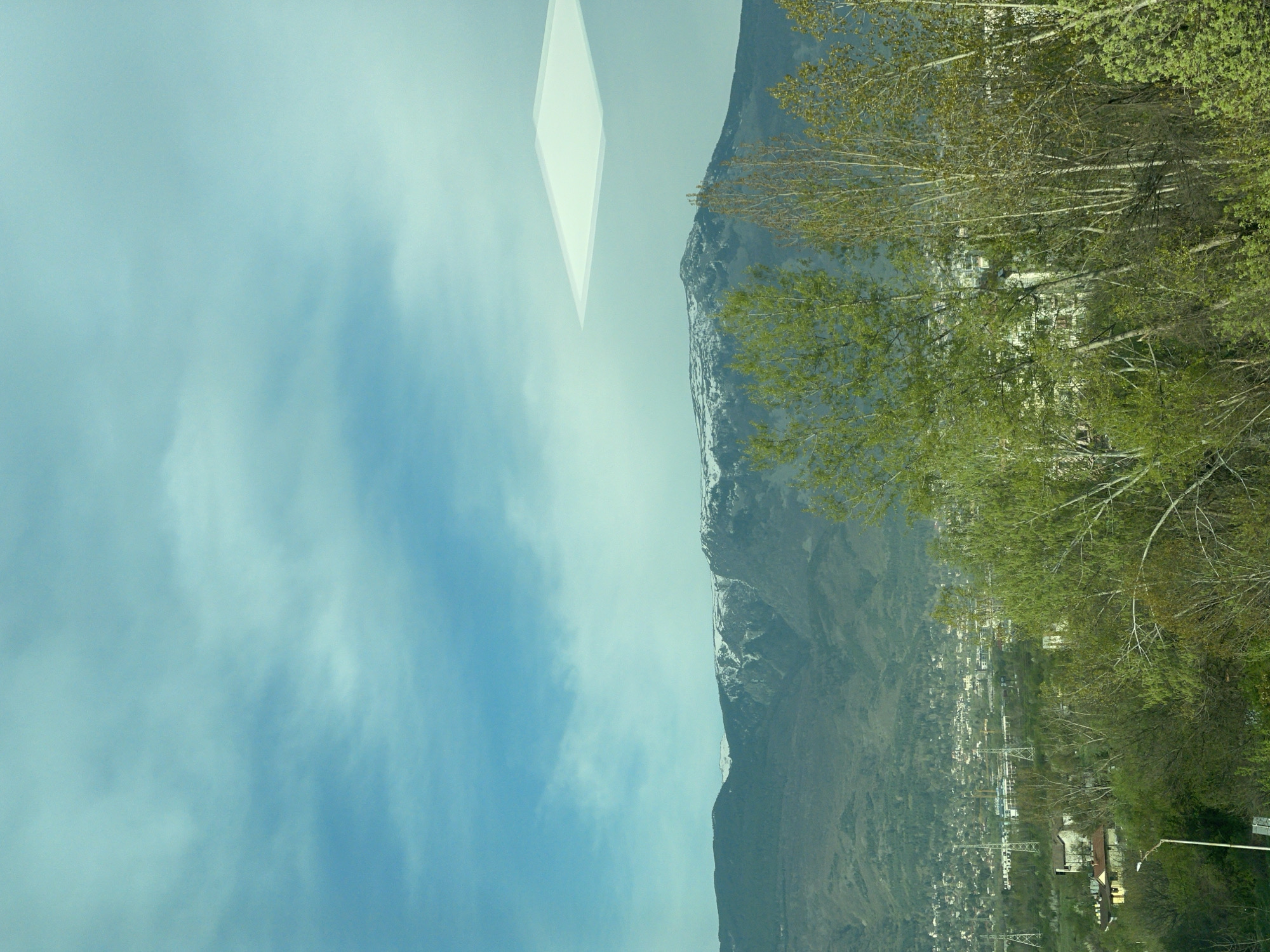 The image displays a landscape featuring a snow-capped mountain range under a partly cloudy blue sky. An urban settlement or town nestles at the base of the mountain. A cluster of trees is visible in the foreground. A kite-shaped, translucent object is visible in the top center of the image. The image's orientation is sideways. The location in the city of Sofia, Bulgaria, is not depicted in the image.