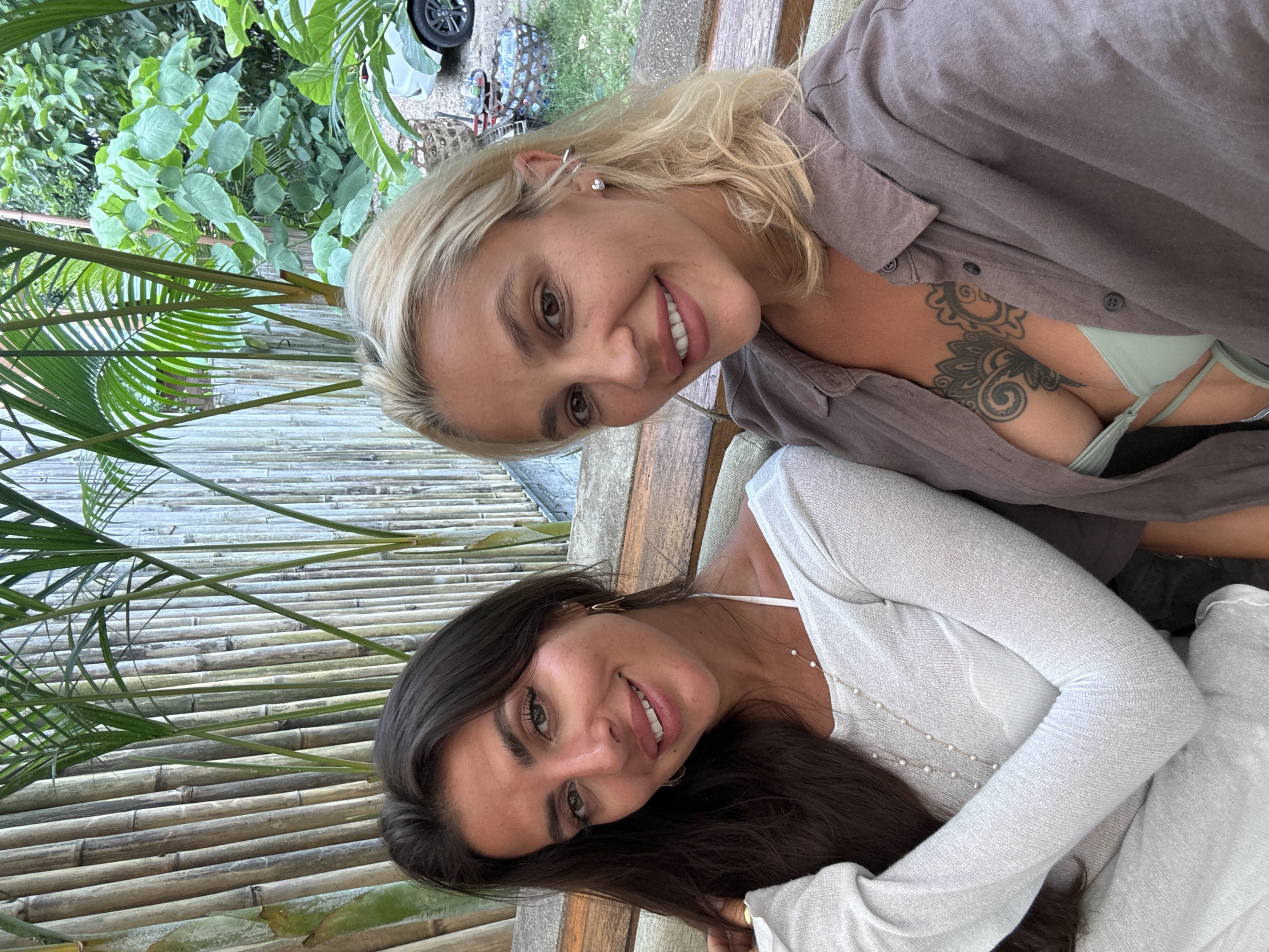 In Kuta Selatan, Indonesia, two women are photographed outdoors, seated on a wooden bench against a backdrop of bamboo stalks and lush foliage. The woman on the left has long, dark hair and is wearing a white, long-sleeved top adorned with a pearl necklace. The woman on the right has blonde hair and is wearing a grey open shirt revealing a tattoo on her chest and a green bikini top. Both women are smiling and looking at the camera, suggesting a candid or posed selfie. The scene appears to be a relaxed, tropical setting, possibly a resort or restaurant given the natural elements and seating arrangement.